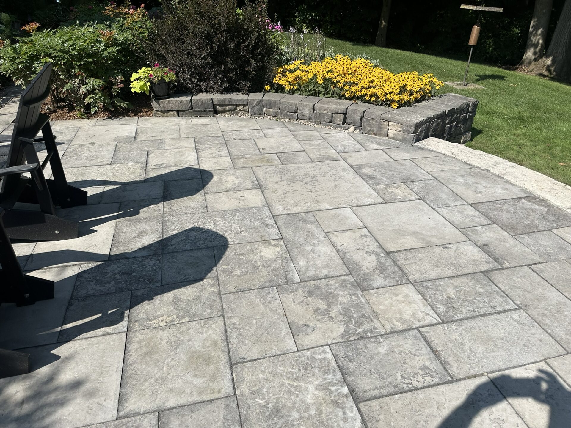 A stone patio with black chairs, surrounded by vibrant yellow flowers and greenery, under bright sunlight, casting distinct shadows on the tiles.