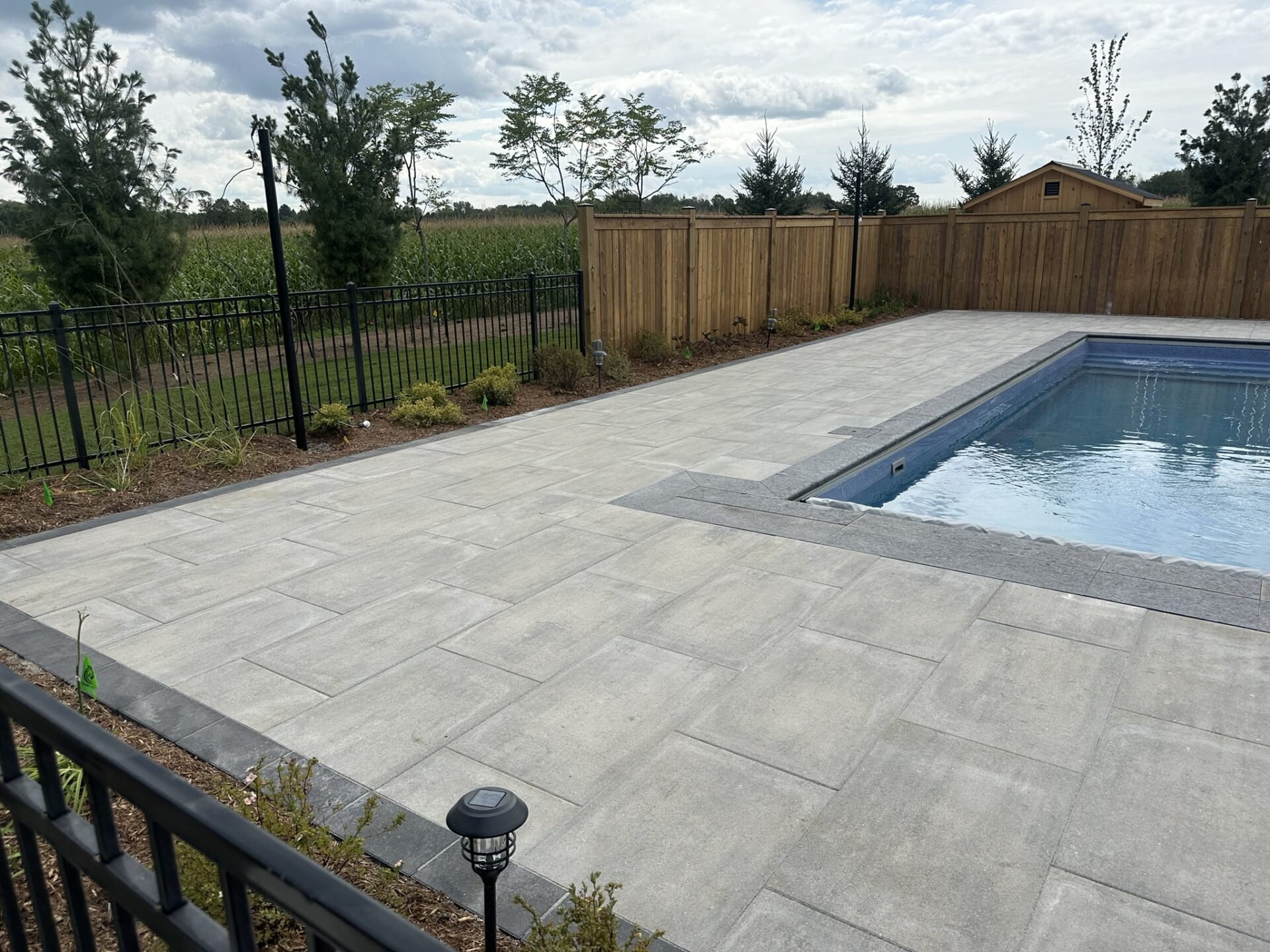 A modern backyard features a rectangular pool, paved patio, fencing, and young trees, set against a rural landscape and cloudy sky.