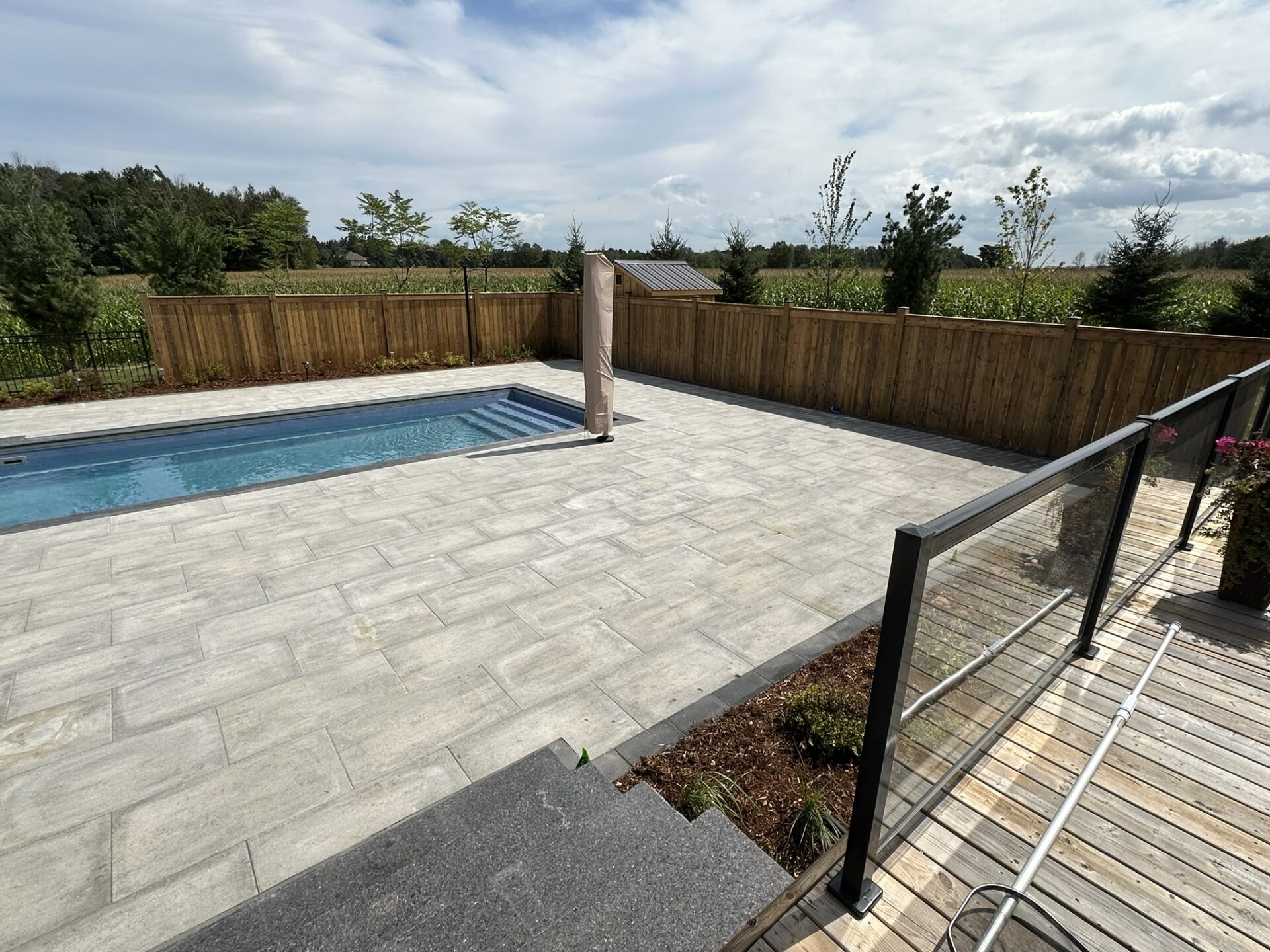 A private backyard features a rectangular pool, stone patio, wooden fence, and glass railing, surrounded by trees under a partly cloudy sky.