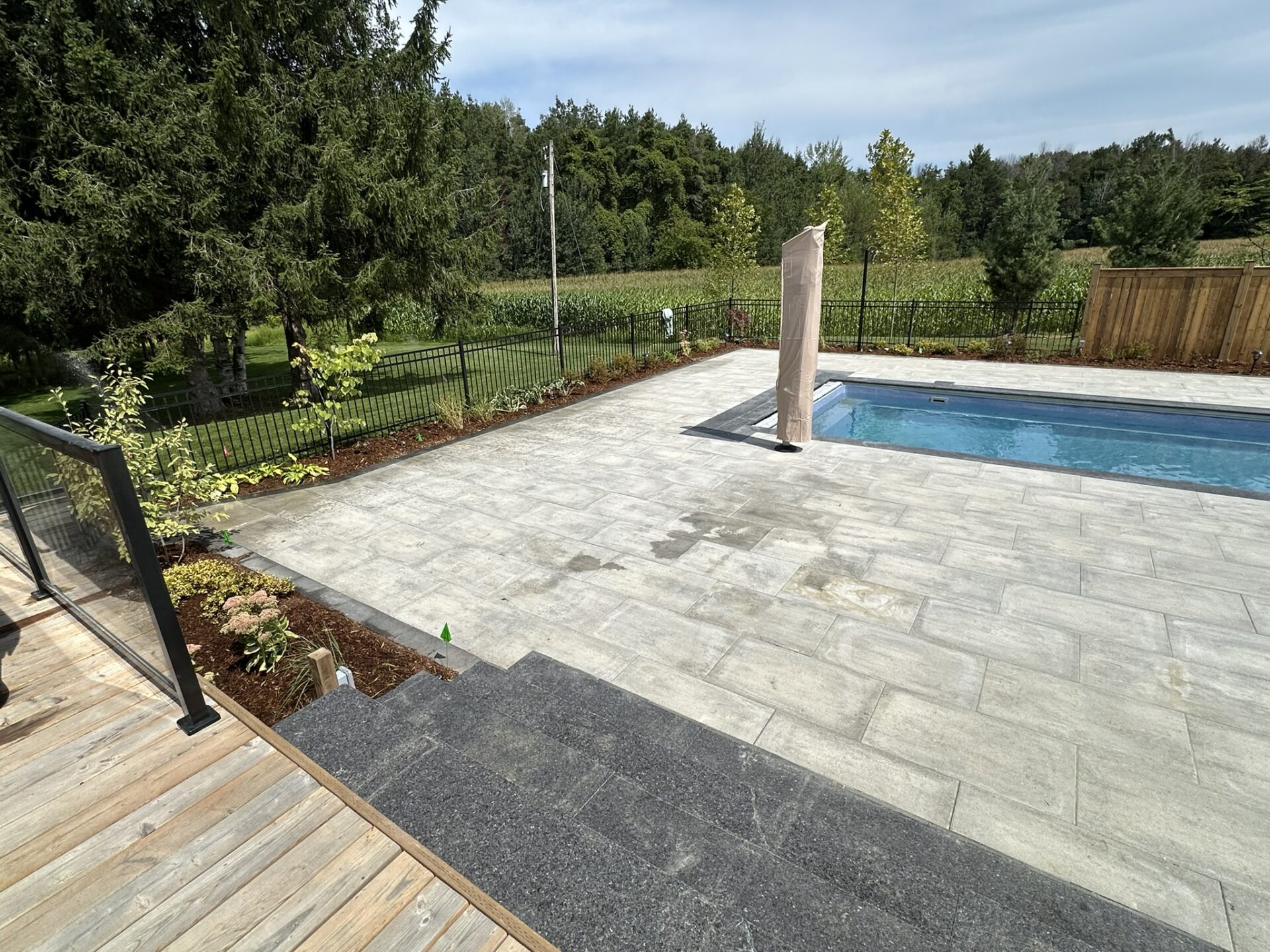 A backyard with a pool, surrounded by trees and a fence, featuring a stone patio and wooden steps for outdoor relaxation.