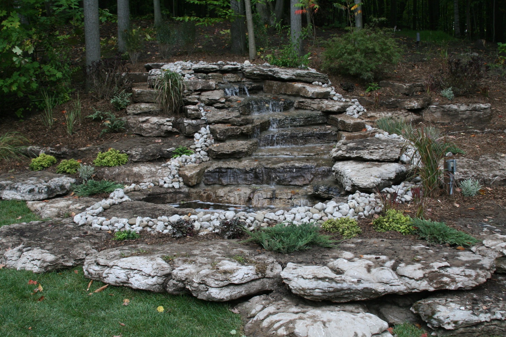 A serene rock waterfall feature in a landscaped garden, surrounded by greenery and trees, creating a natural and tranquil setting.