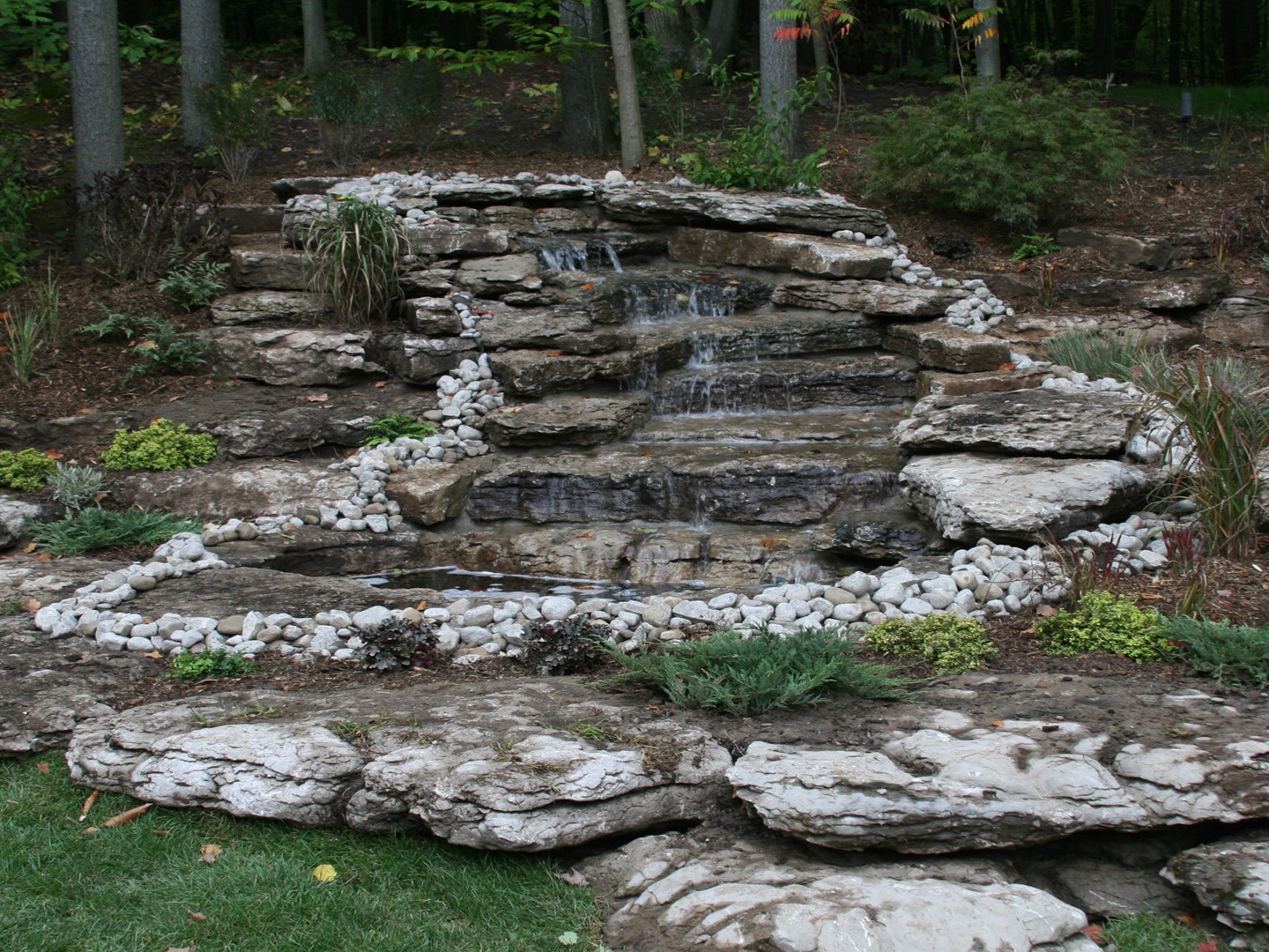 A tiered rock waterfall in a garden setting surrounded by green foliage and trees, creating a tranquil, natural landscape.