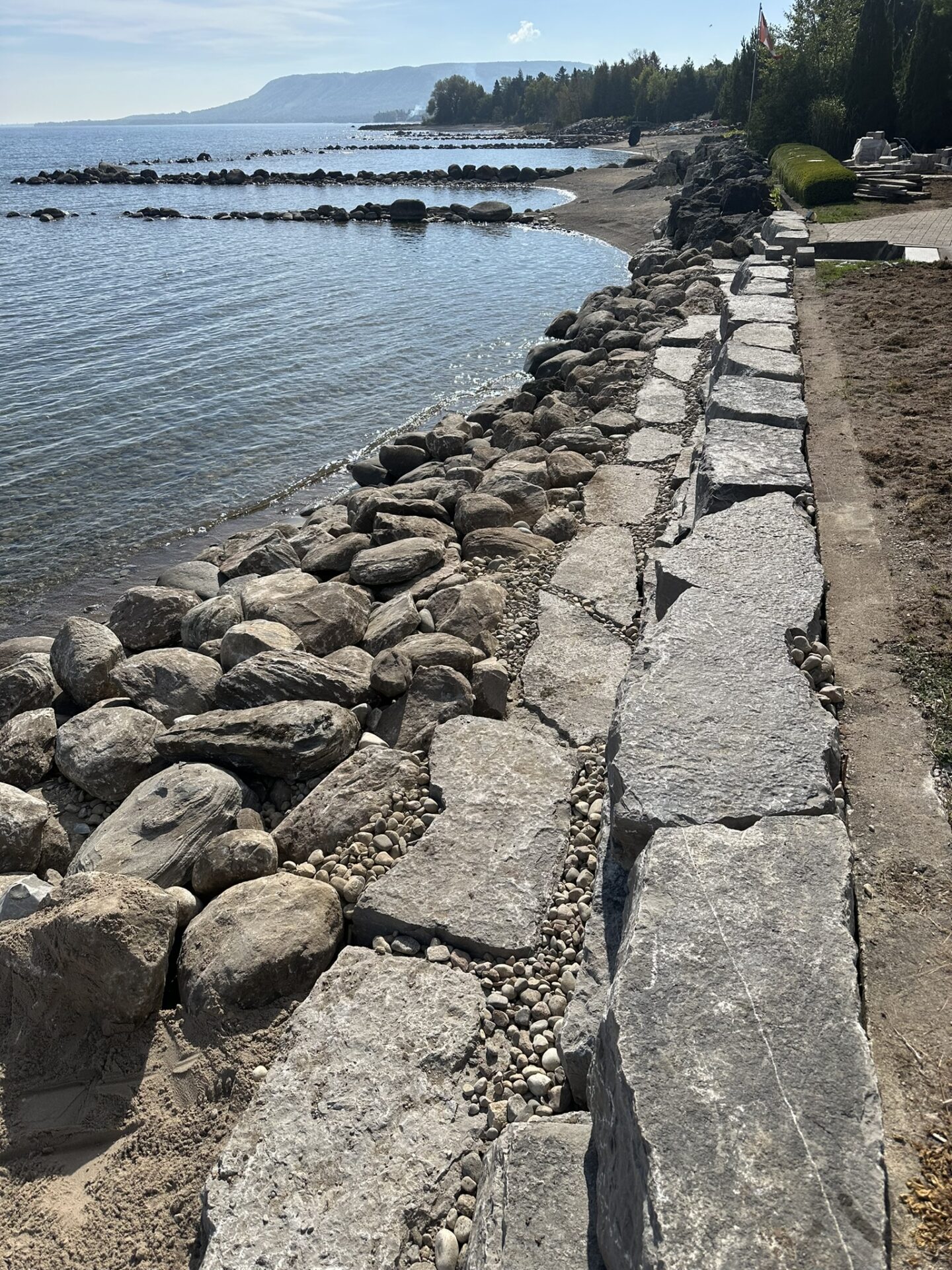 Rocky shoreline with a stone retaining wall alongside a calm body of water, bordered by trees under a clear, sunny sky.