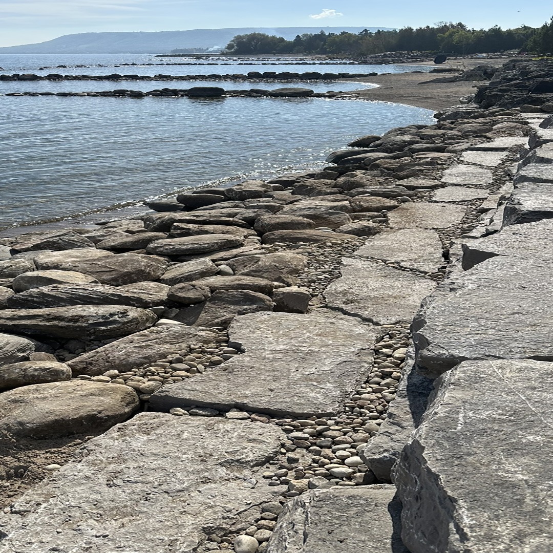 Rocky shoreline with flat stones and pebbles leading to a calm lake. Trees and hills are visible under a clear sky.