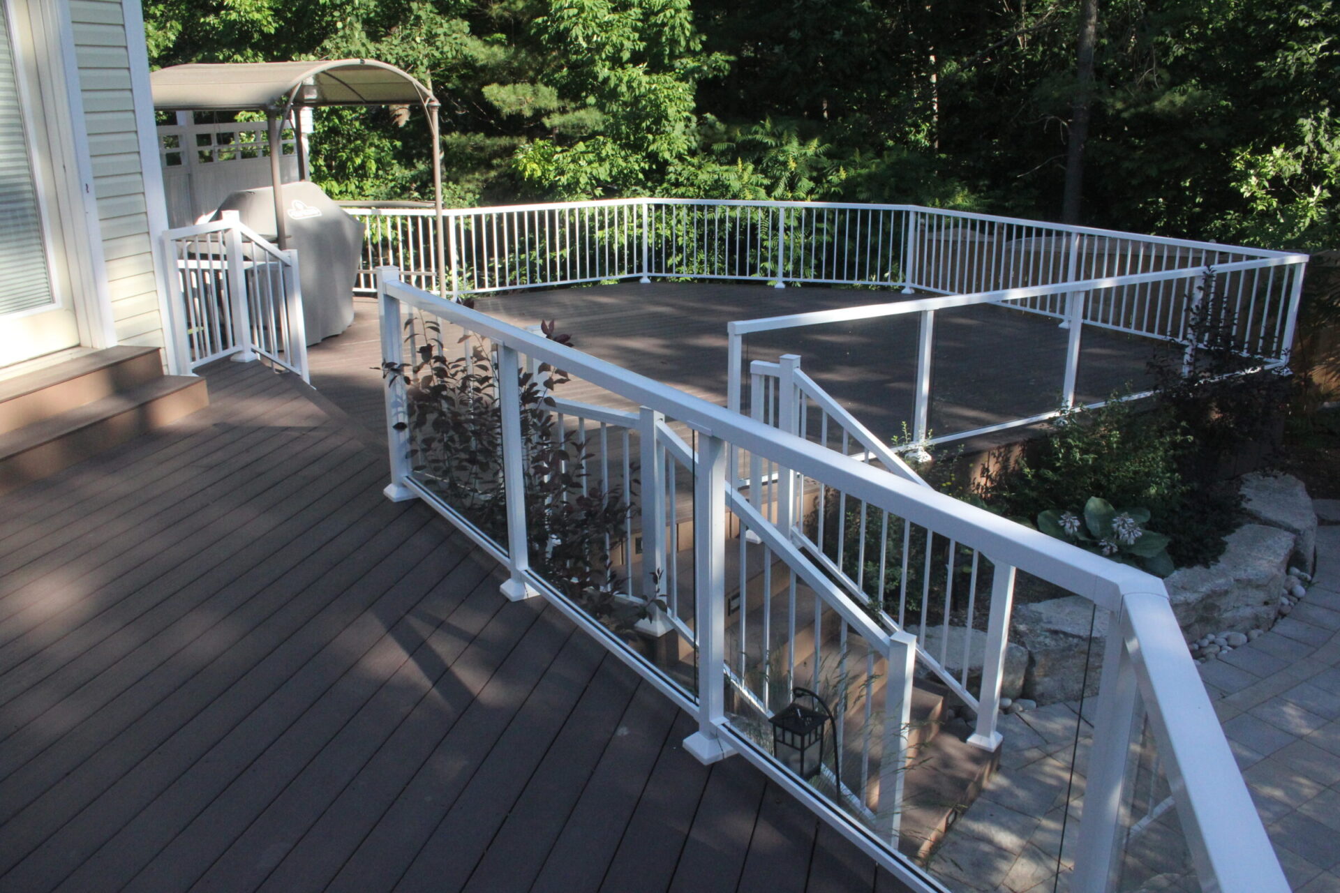 A spacious wooden deck with white railing, outdoor grill, surrounded by lush greenery, leading to a garden area with stone pathway.