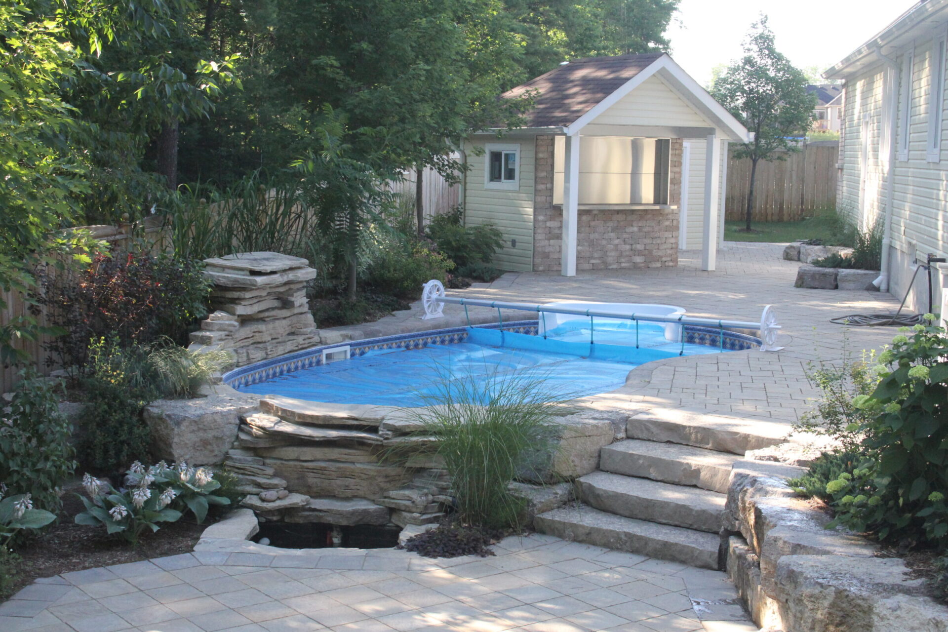 A backyard scene featuring a small pool with a cover, surrounded by stone landscaping and greenery. There is a small pool house nearby.