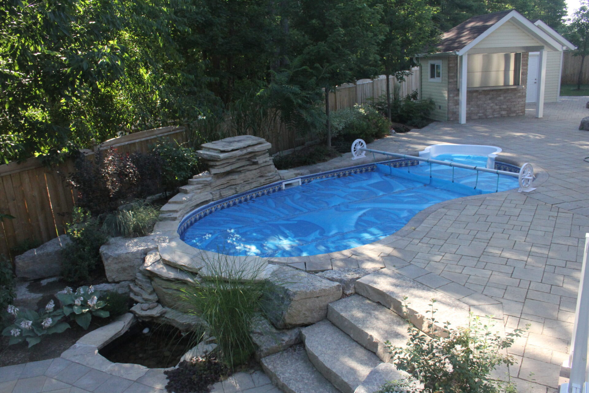 Backyard view featuring a small, curvy blue swimming pool surrounded by stone landscaping and trees, with a stone patio and a small pool house.