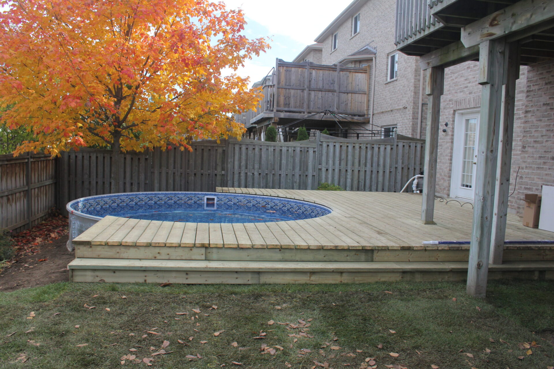 A backyard scene with a wooden deck, above-ground pool, vibrant orange tree, and fenced surroundings near brick residential buildings, under a clear sky.