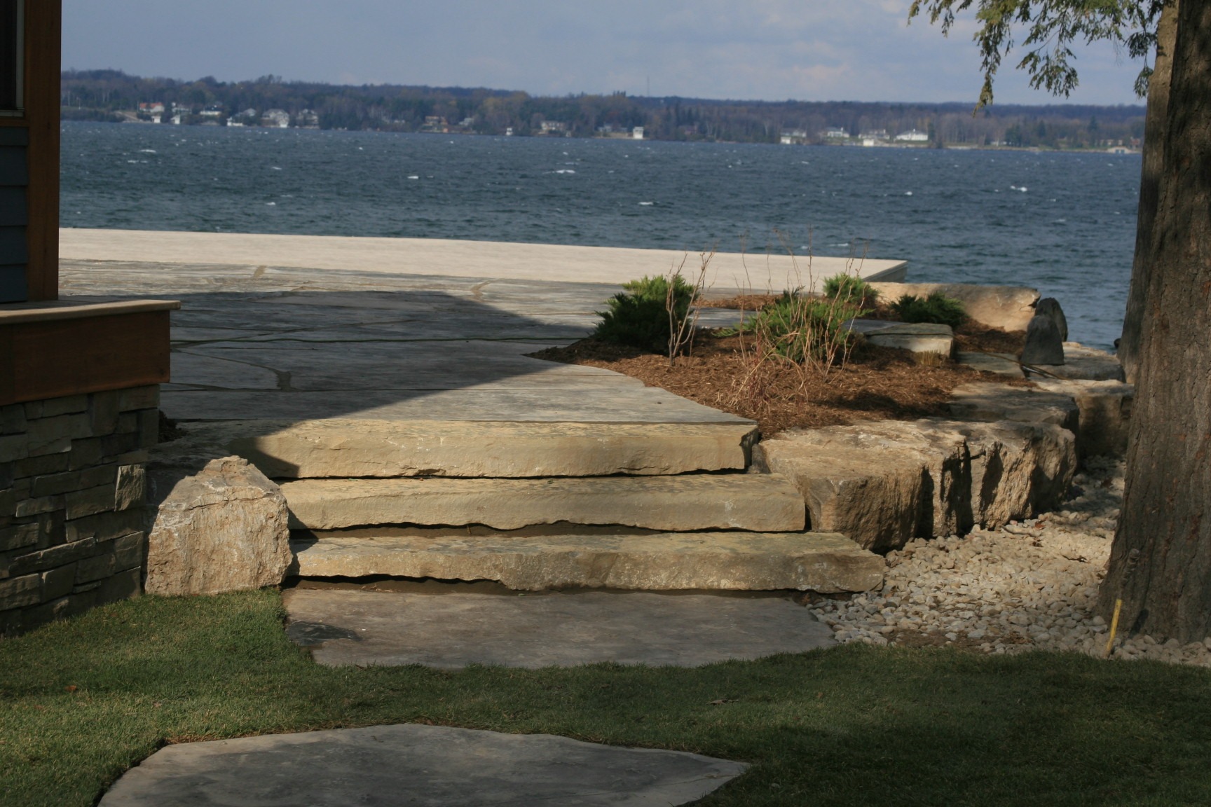 Stone steps lead to a lakeside patio area with a scenic view of the water and distant shoreline under a partly cloudy sky.