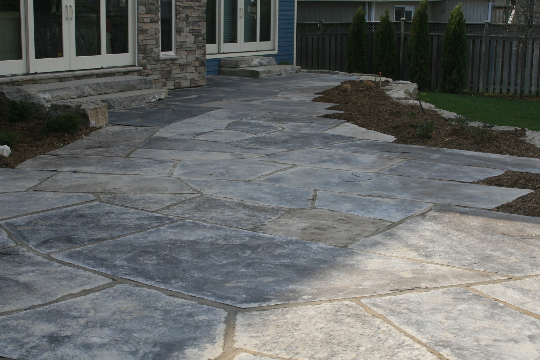 A stone patio featuring irregular flagstone design, bordered by a garden bed and adjacent to a contemporary house with large windows and wooden fence.