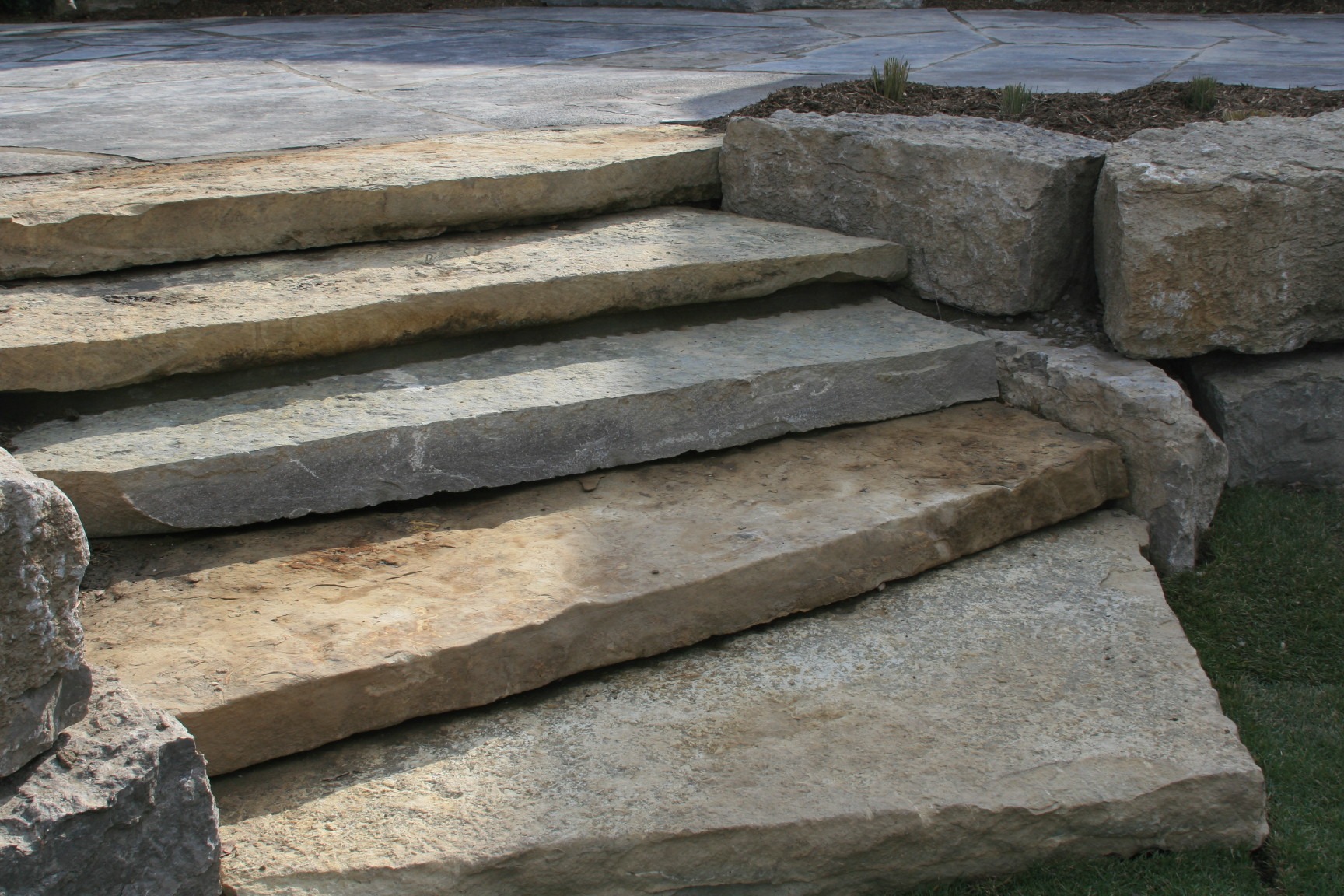 Stone steps leading to a patio area, surrounded by grass and rocks, creating a natural, rustic outdoor setting. No identifiable landmarks present.