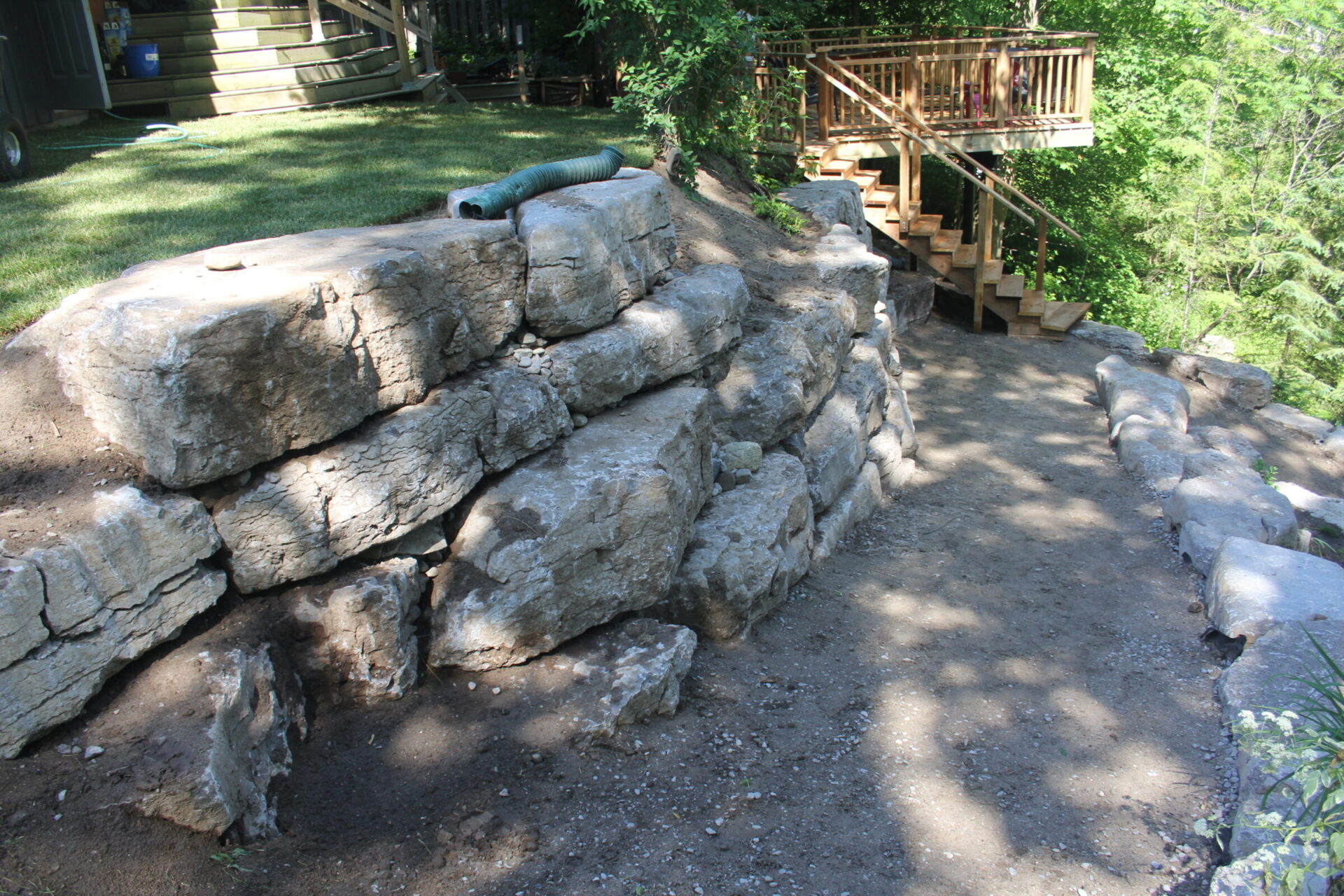 Stone retaining wall with wooden stairs leads to an elevated deck surrounded by lush greenery, creating a peaceful outdoor setting.