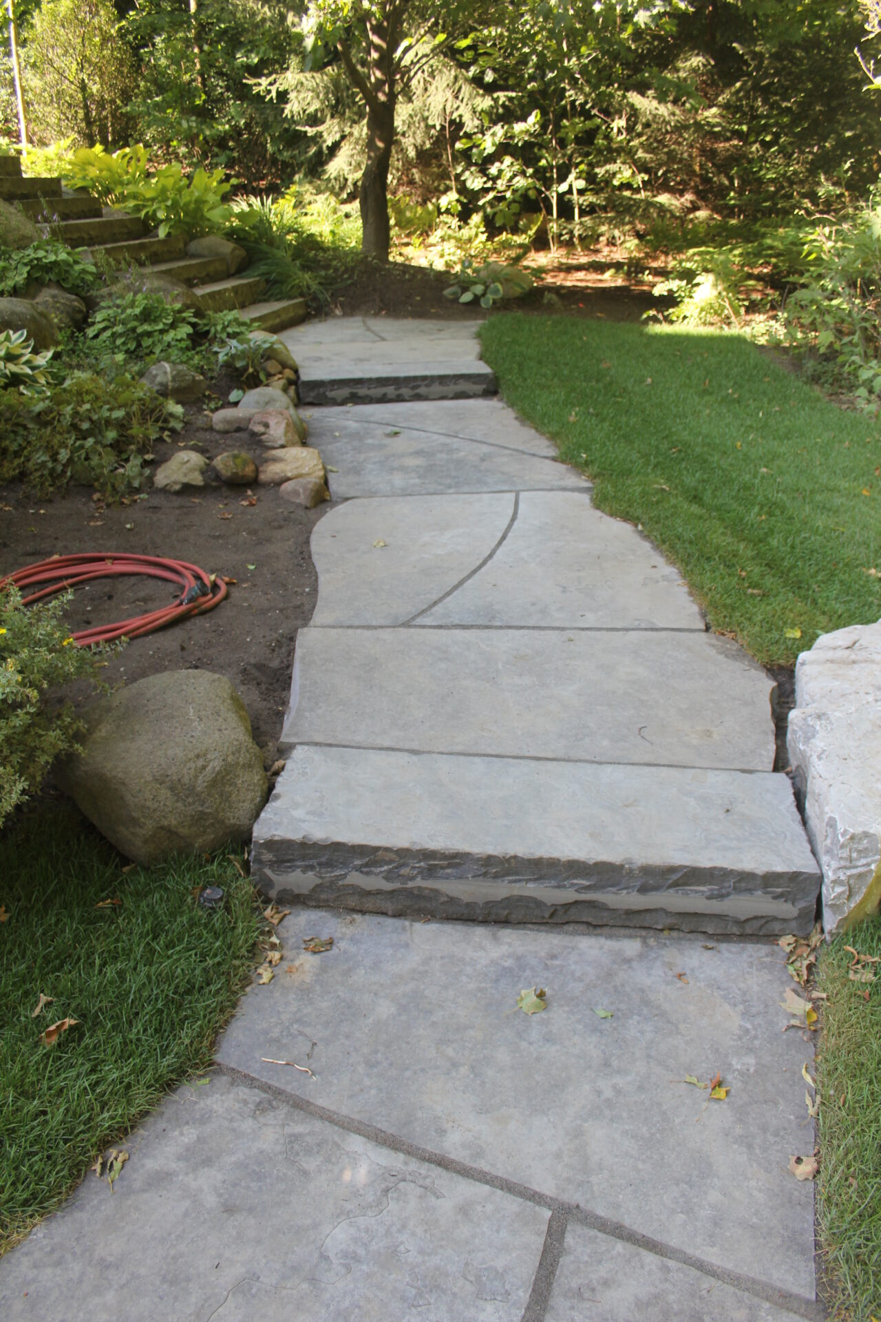 A stone pathway curves through a lush garden, bordered by grass and shrubs, with dappled sunlight filtering through the trees overhead.