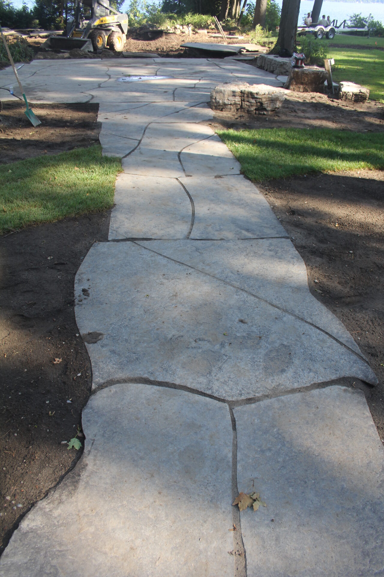 A winding stone pathway under construction is surrounded by grass and trees. A person is sitting on a wooden stump nearby.