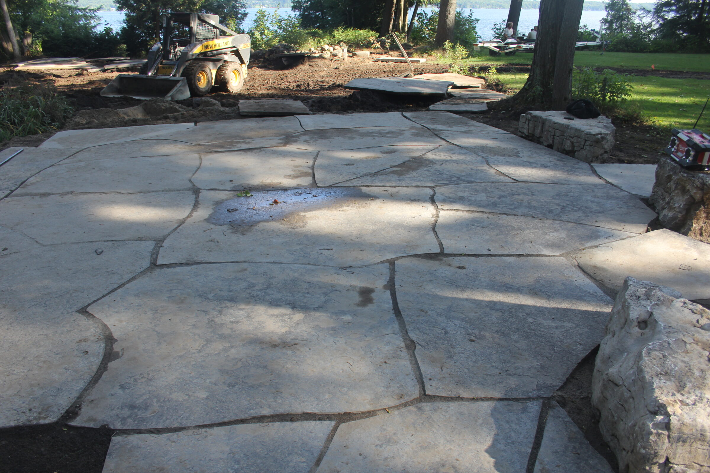 Stone patio under construction in a wooded area. Equipment and loose stone visible. Trees and distant water view in the background.