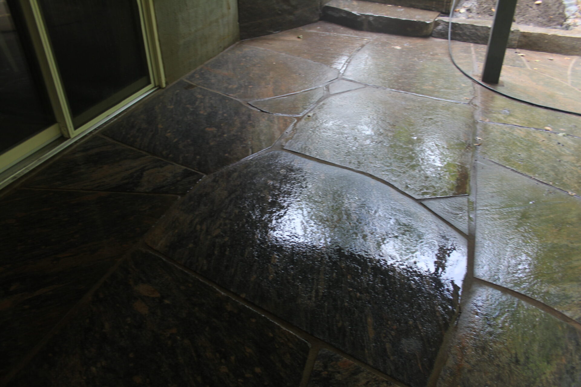 A wet stone patio with natural, irregularly shaped tiles. Reflections on the surface enhance the visual texture and ambiance.