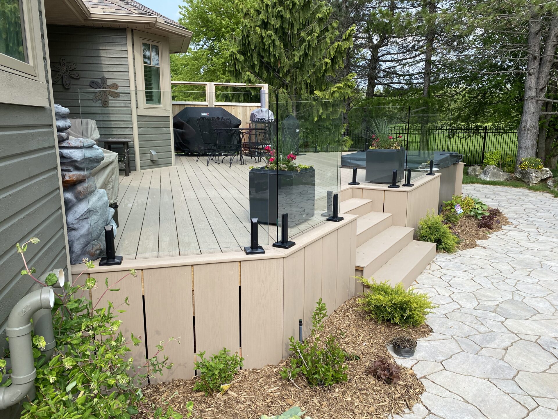 Elevated wooden deck with glass railing overlooks a stone pathway. Patio furniture and barbecue grill visible, surrounded by trees and shrubs.