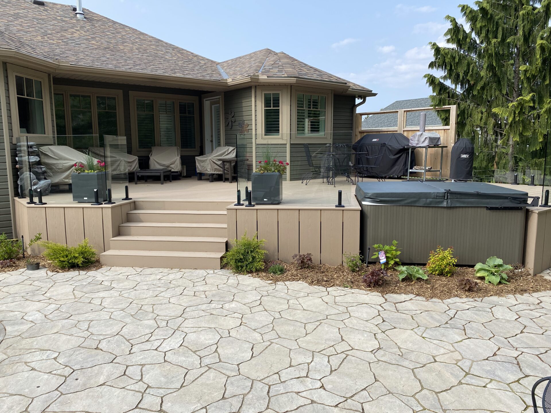 A modern backyard patio with steps, seating, and a covered hot tub. Stone pavement and greenery enhance the cozy outdoor space.