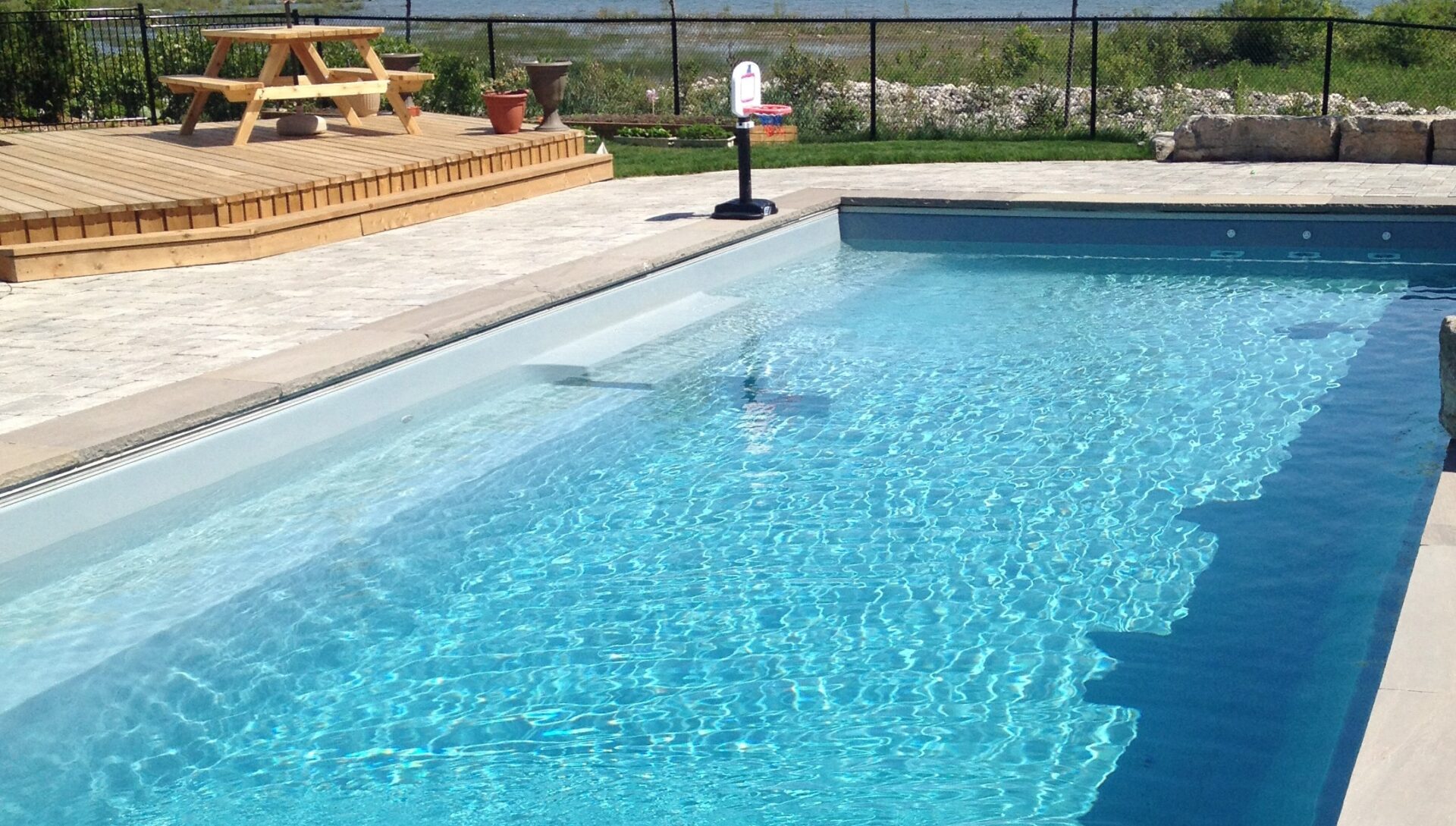 A rectangular swimming pool with clear blue water bordered by a paved deck. Nearby is a wooden picnic table. Grass and stones in the background.