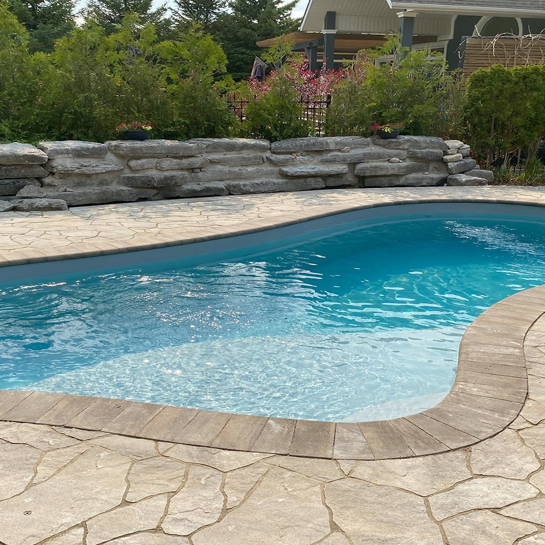 A backyard pool surrounded by stone pavement and lush greenery on a sunny day, creating a serene and inviting atmosphere.