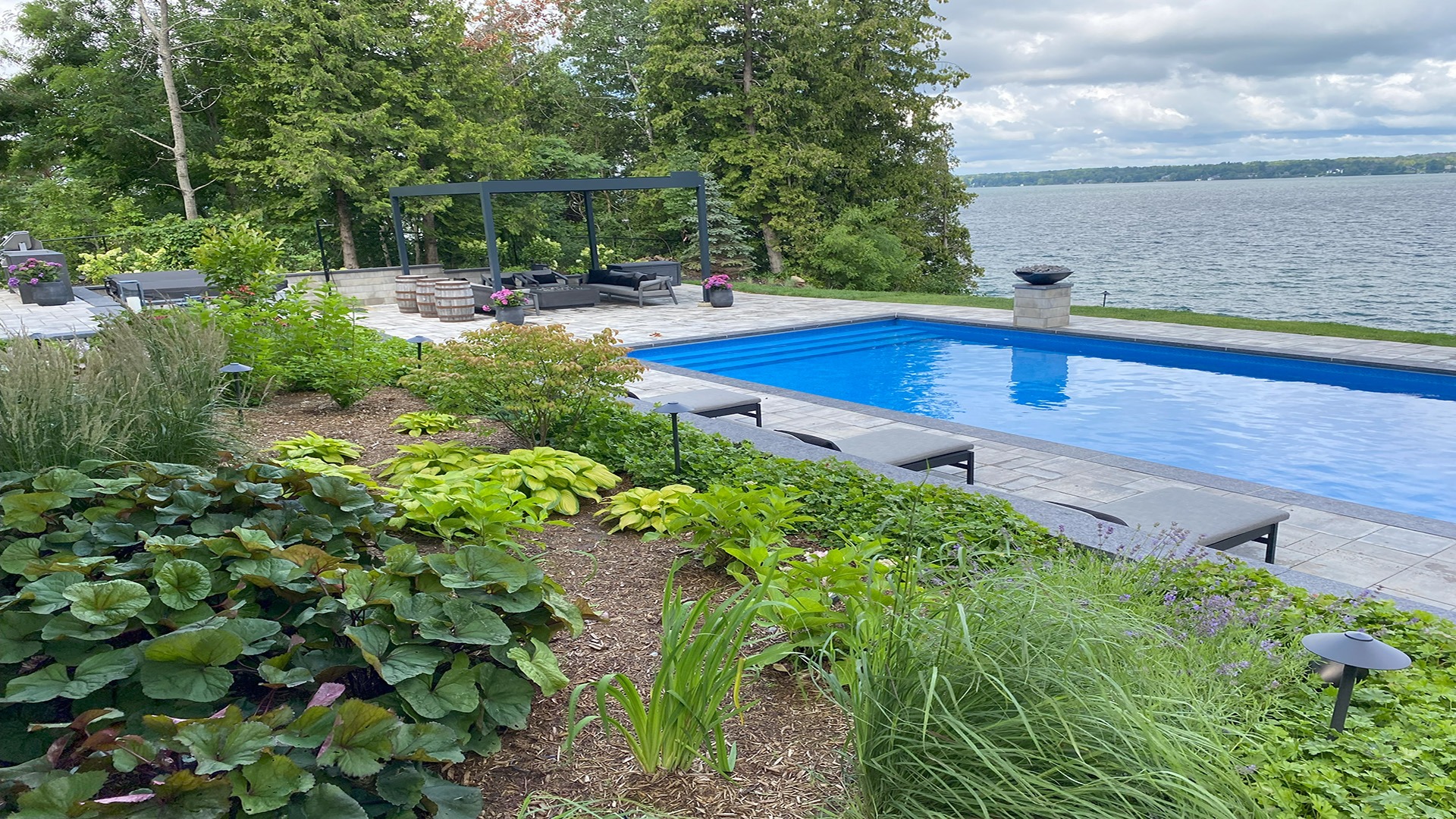 A serene lakeside scene with a pool, lush greenery, lounge chairs, and a seating area, surrounded by tall trees and water views.