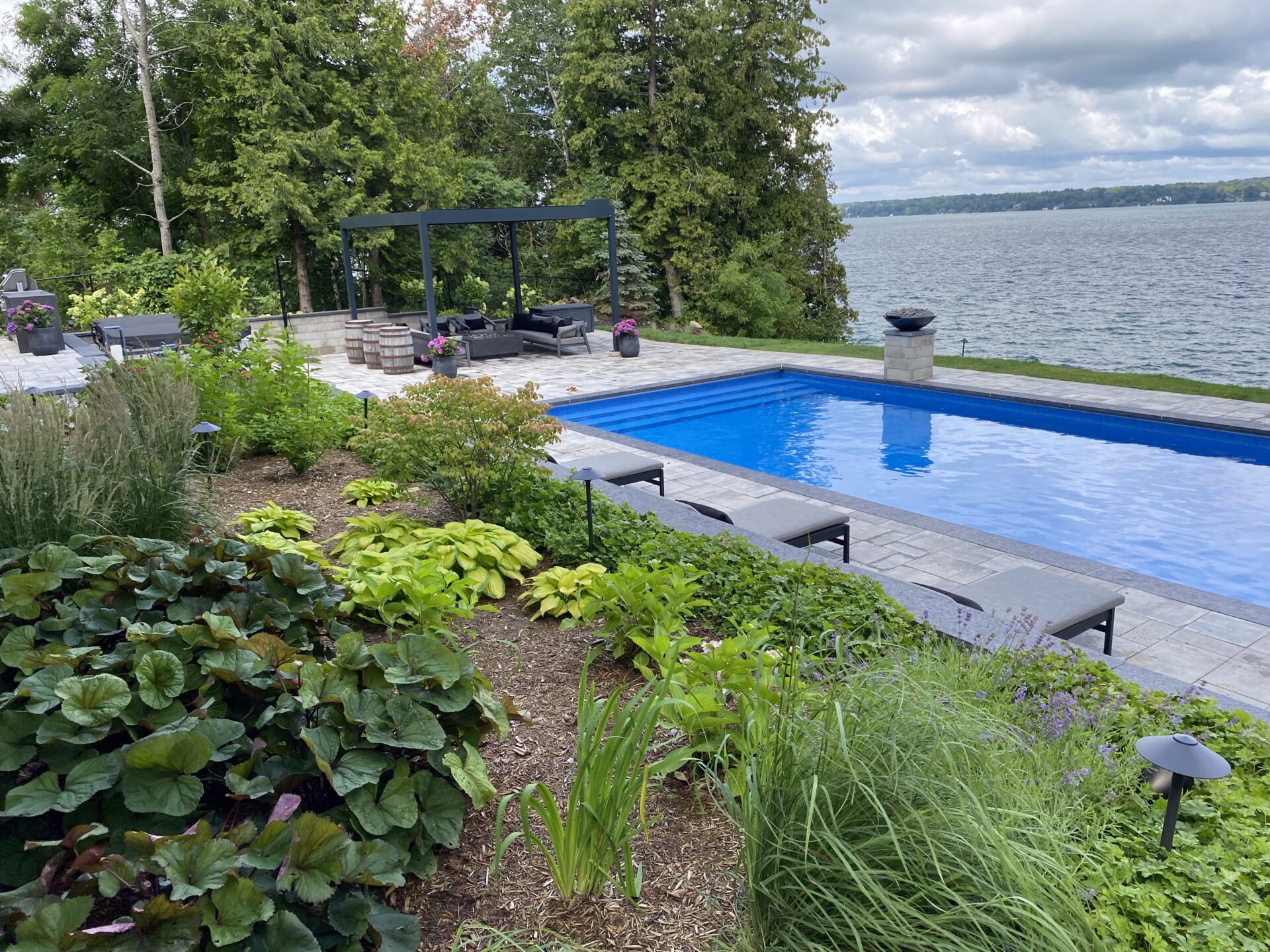 A serene lakeside scene features a blue pool, lush greenery, lounge chairs, and a shaded patio area under a mostly cloudy sky.