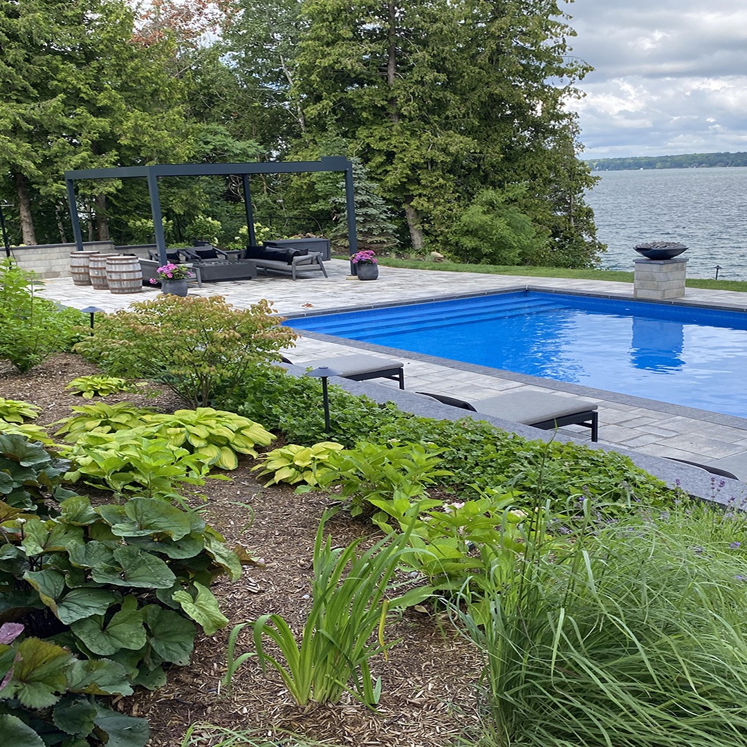 A backyard features a blue swimming pool, outdoor seating under a pergola, lush greenery, and overlooks a large body of water.