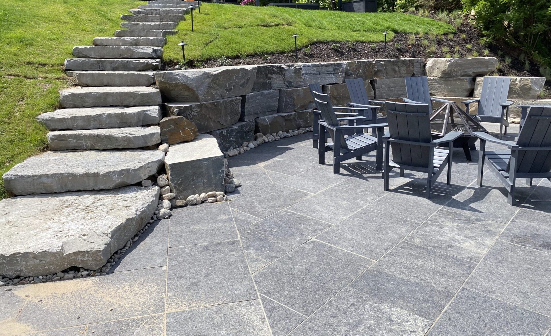 Stone stairs and patio with wooden chairs and table. Surrounded by grass and rocks, creating a serene outdoor seating area.