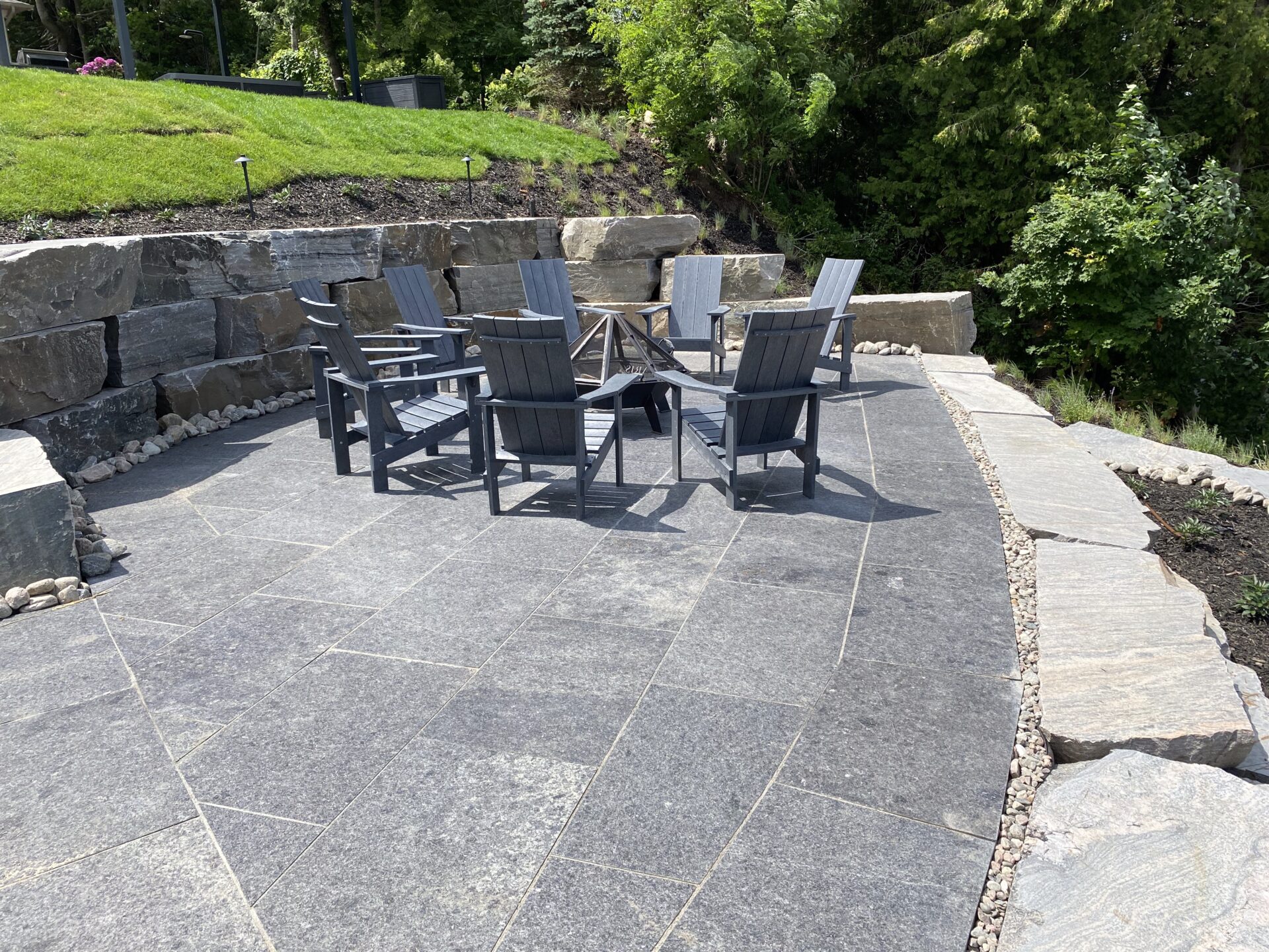 A stone patio with eight wooden chairs arranged around a small fire pit, surrounded by greenery and retaining walls on a sunny day.