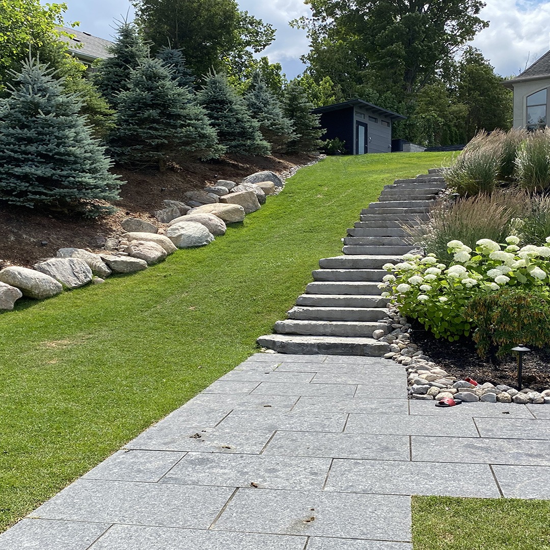 Stone pathway and steps lead through a landscaped garden with lush greenery, small trees, and white flowers. A shed is visible in the background.