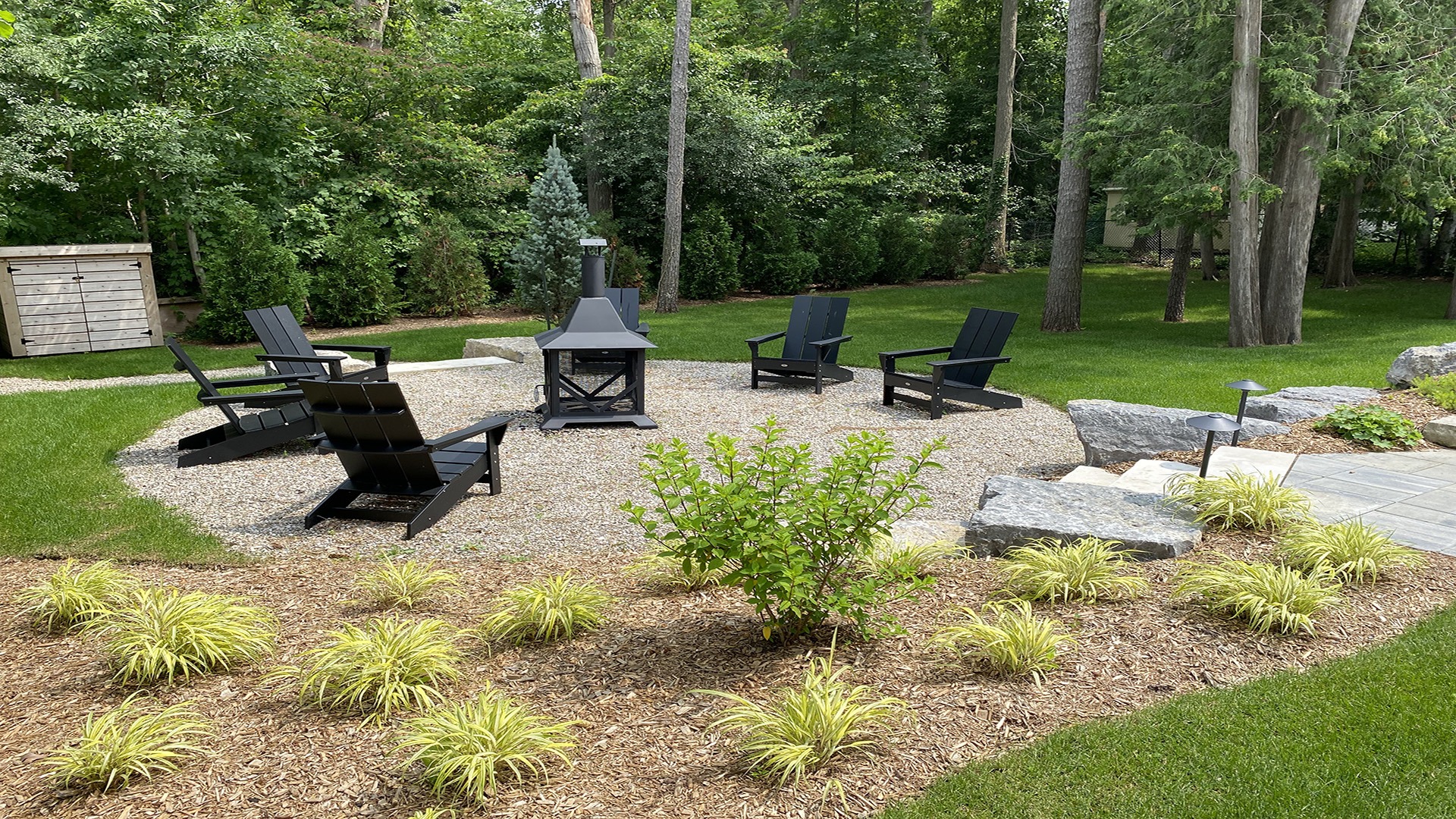 A quiet backyard setting features black chairs around a modern firepit, surrounded by lush greenery, mulch, and a stone pathway.