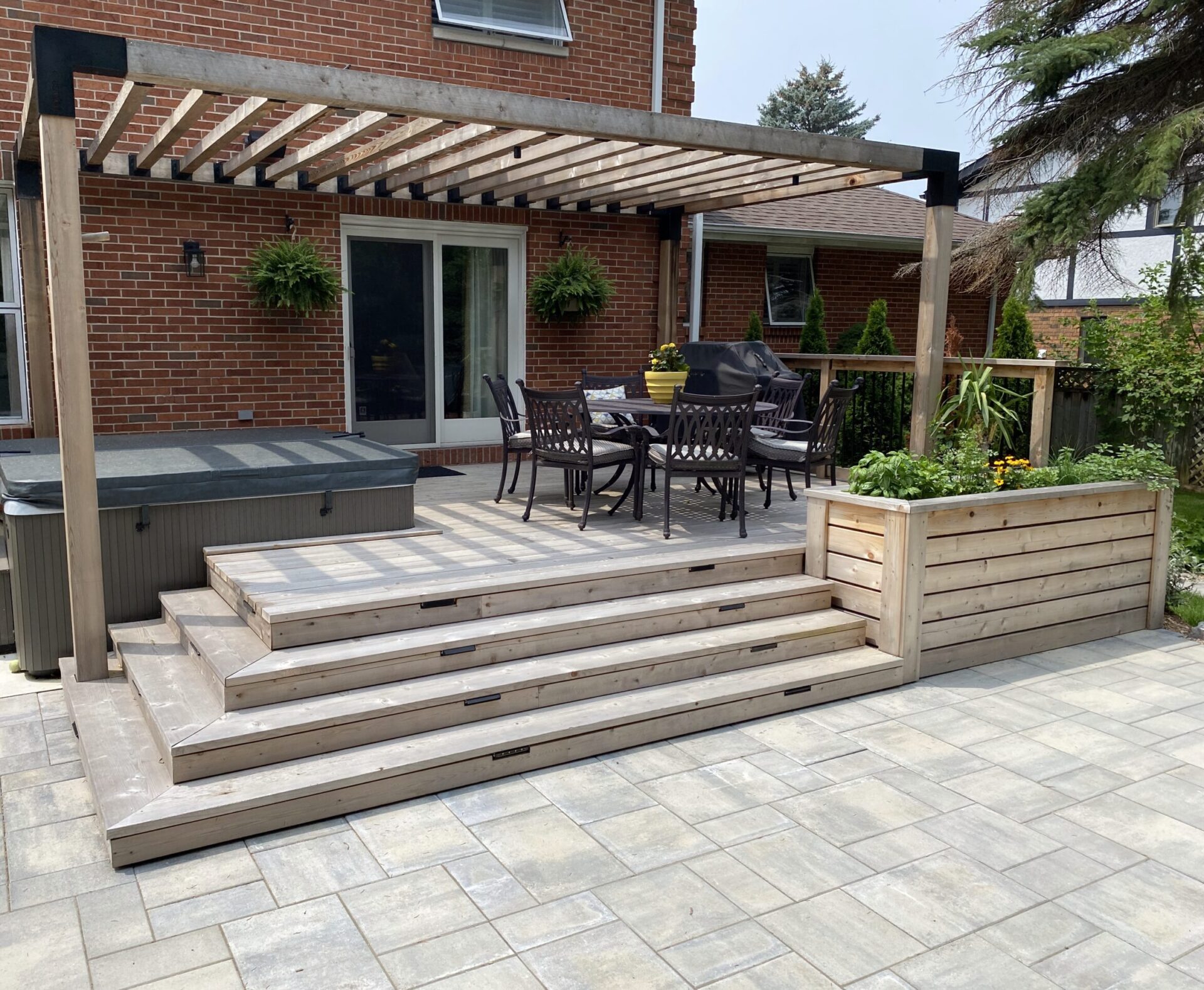 Wooden deck with pergola features outdoor dining set, lush planter, and adjacent hot tub. Brick house serves as a backdrop, patio paved.