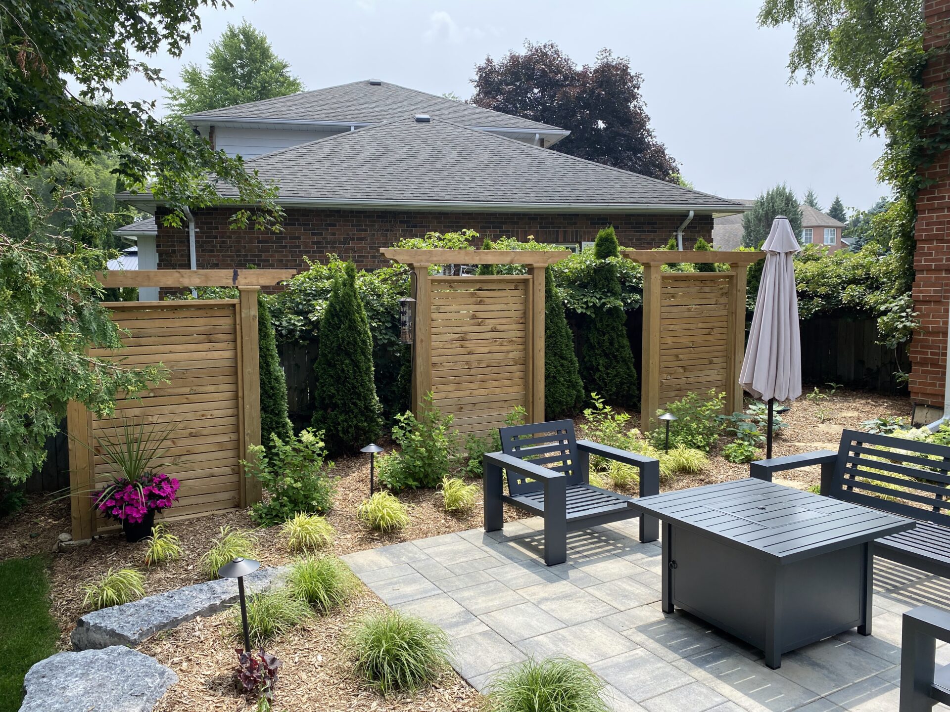 A private backyard features a patio with outdoor furniture, wooden privacy screens, various plants, and a closed umbrella, all under daylight.