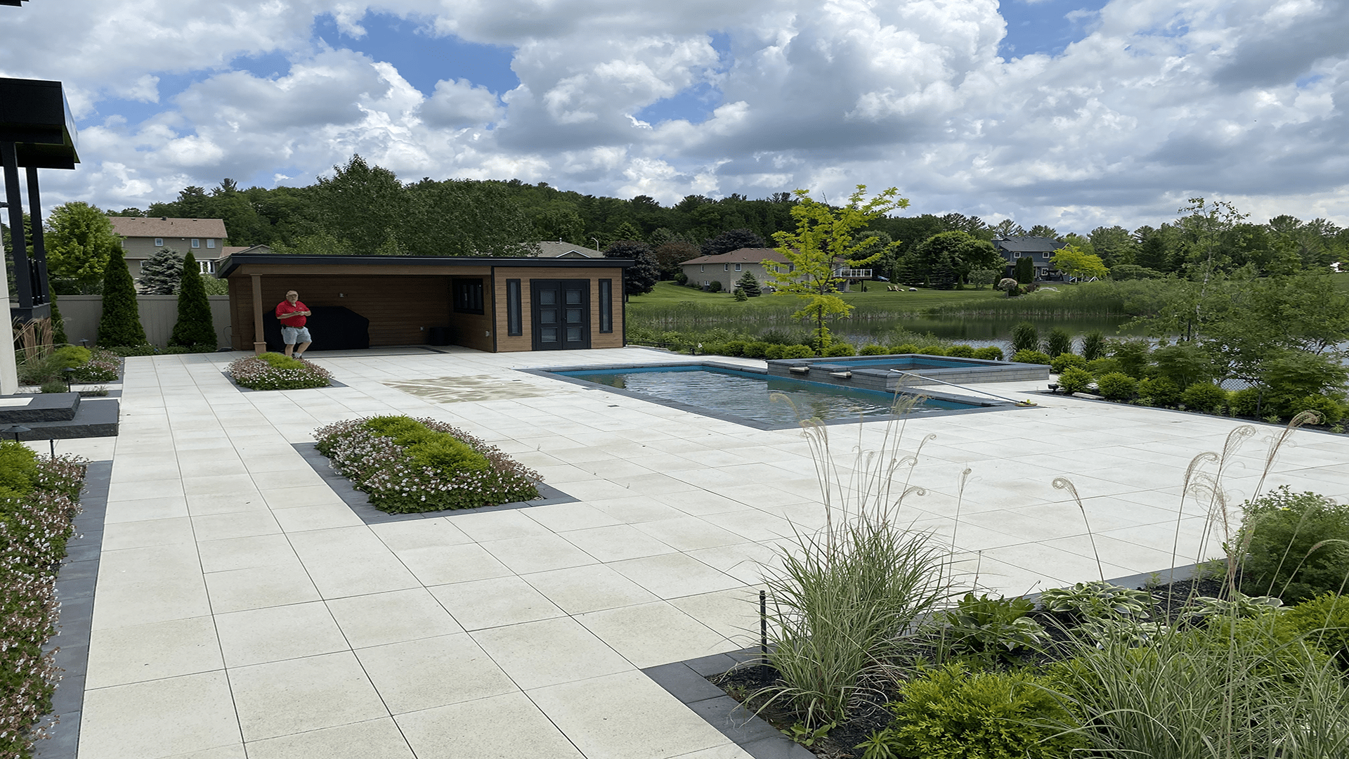 A spacious patio with a modern pool and pool house. A person sits by the building, surrounded by greenery and vast sky.