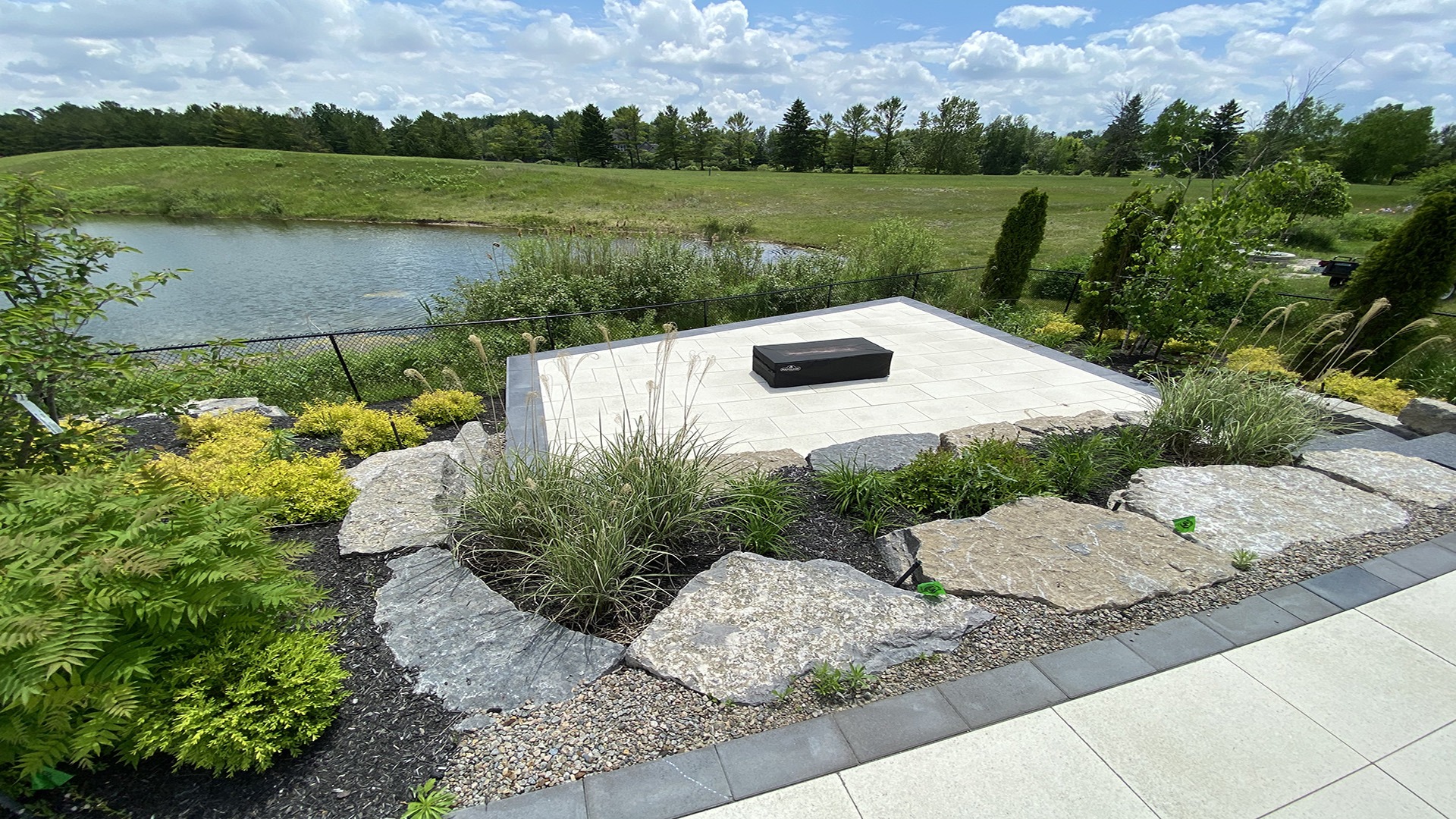 A serene outdoor area features a stone platform surrounded by landscaped greenery, overlooking a tranquil pond under a partly cloudy sky.