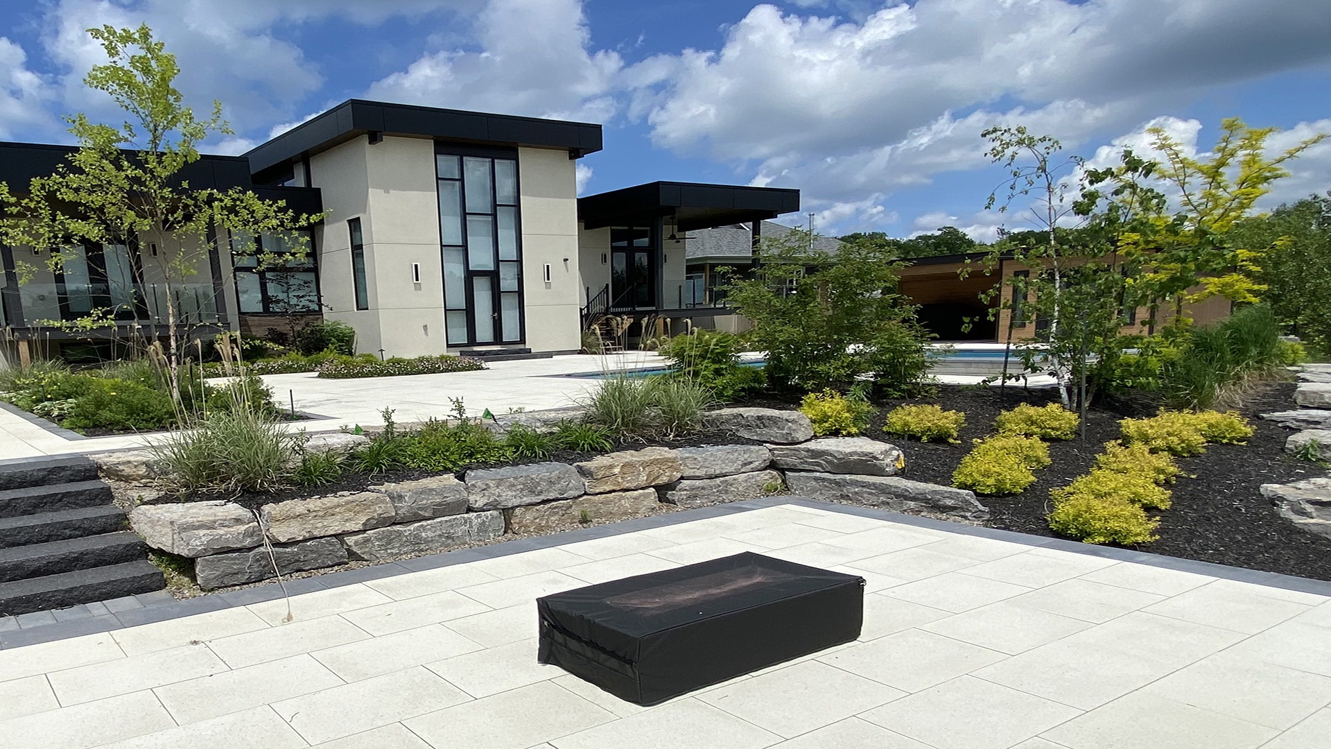 Modern house with geometric design, large windows, and surrounding landscaped garden. Stone pathway and steps lead to the entrance under a blue sky.