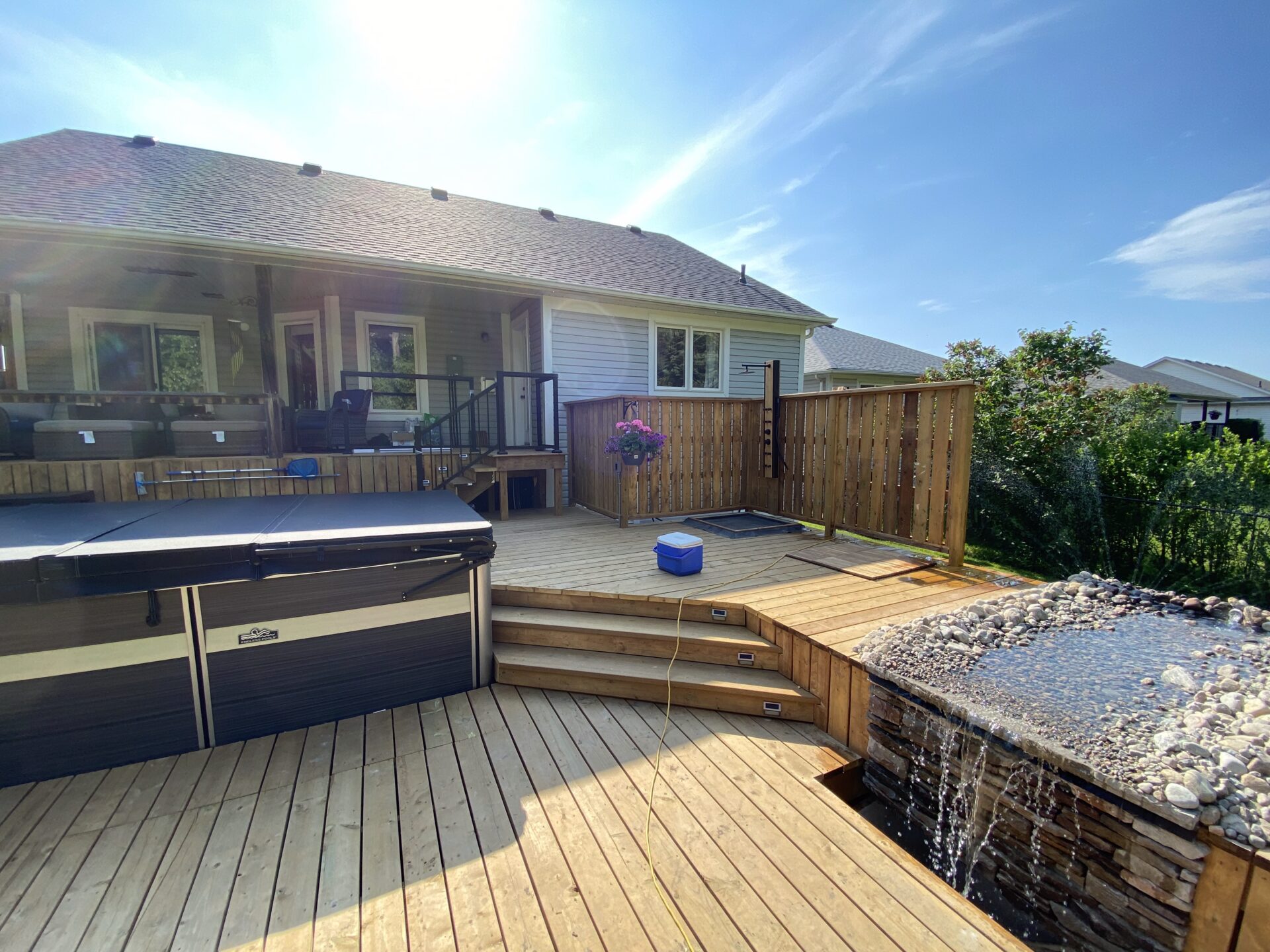 A sunny backyard with wooden decking features a covered patio, hot tub, small waterfall, and hanging plant, creating a relaxing outdoor space.