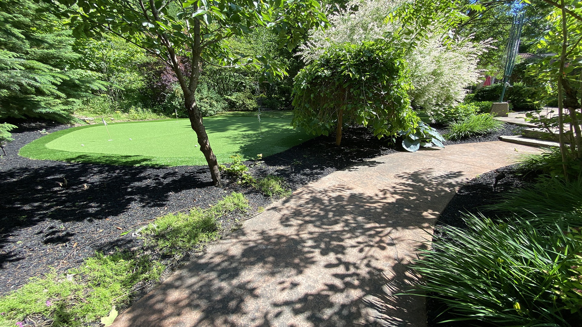 A peaceful garden featuring a winding path, lush greenery, and a small putting green under a clear blue sky.