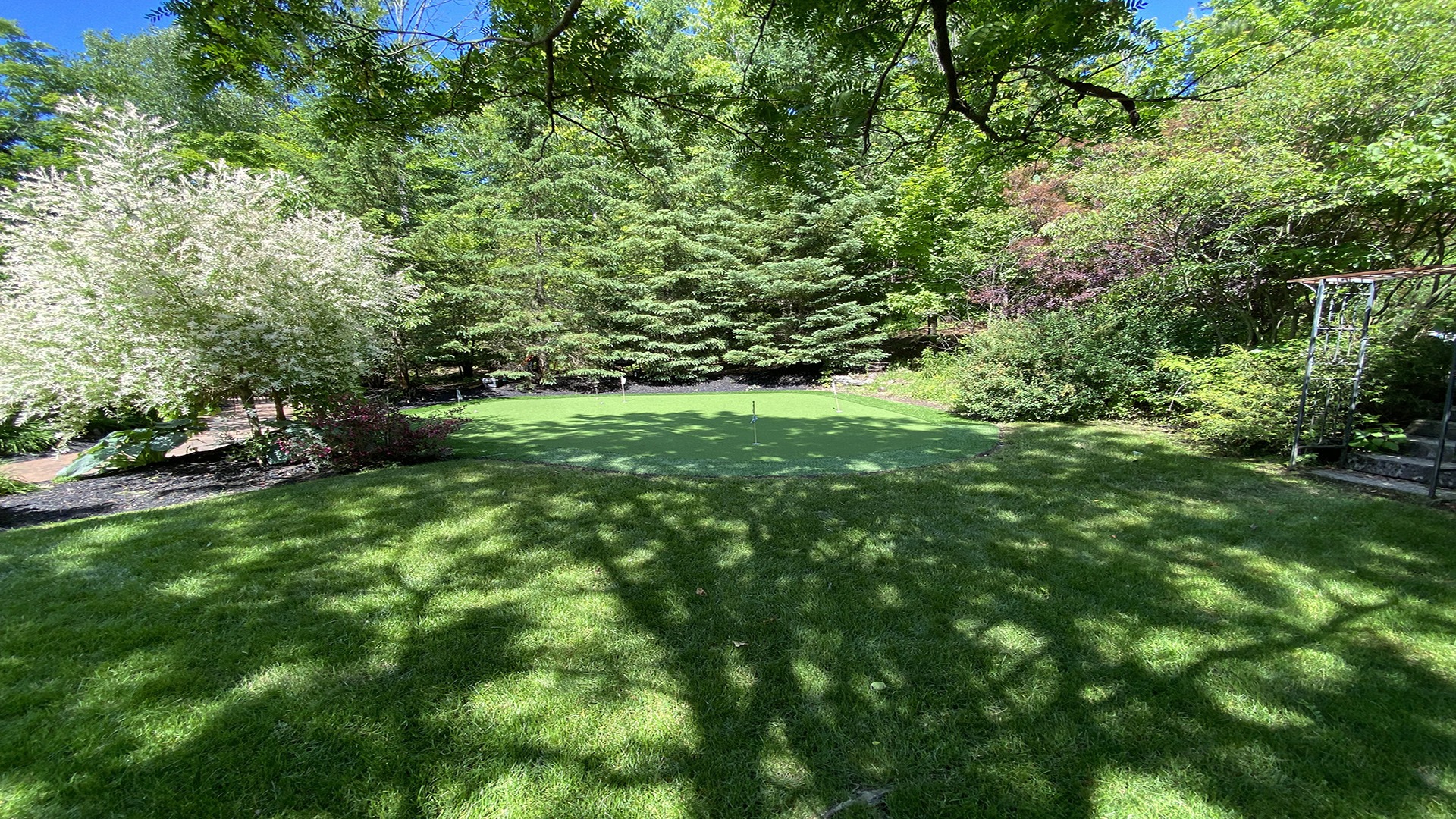 A serene garden scene featuring a lush green lawn, surrounded by various trees and bushes under a clear blue sky.