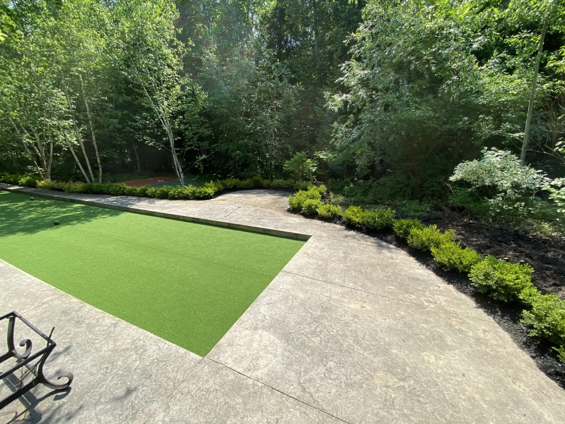 A landscaped garden with a green artificial turf strip, surrounded by trees and shrubs. Stone pathways and a decorative bench are visible.