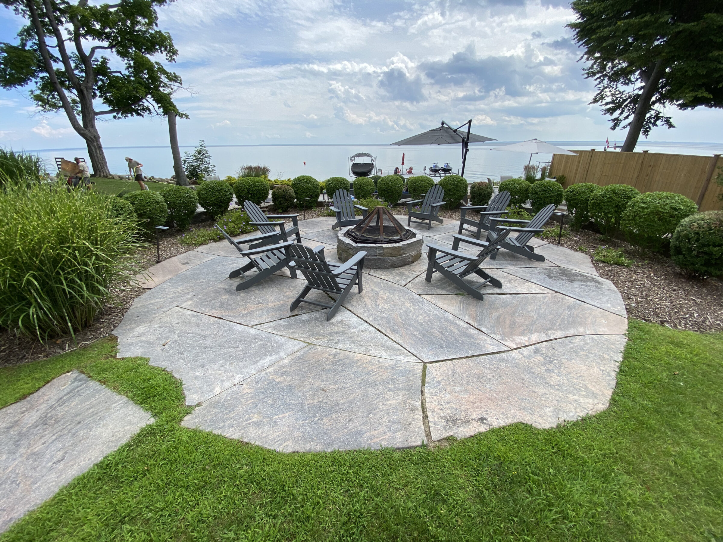 Circular seating area with Adirondack chairs around a fire pit, overlooking a lake. Surrounded by greenery and a stone pathway.
