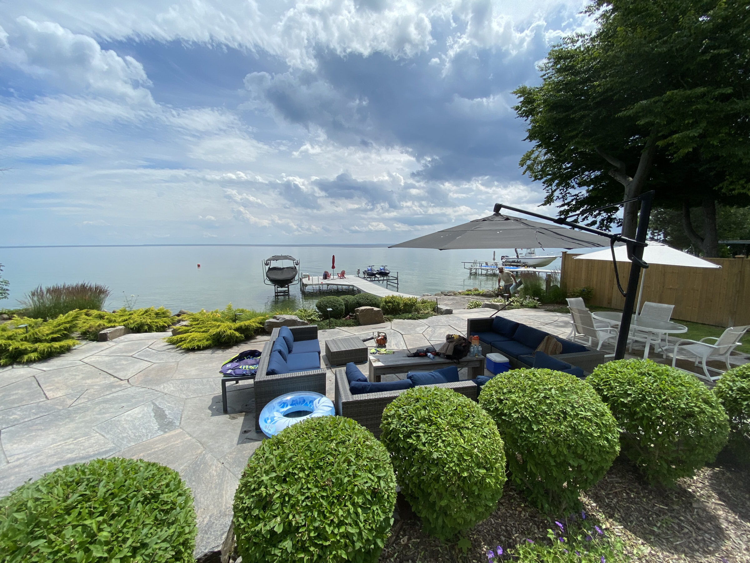 A serene lakeside patio with comfortable seating, lush greenery, and a large umbrella, overlooking a dock and calm waters under a cloudy sky.