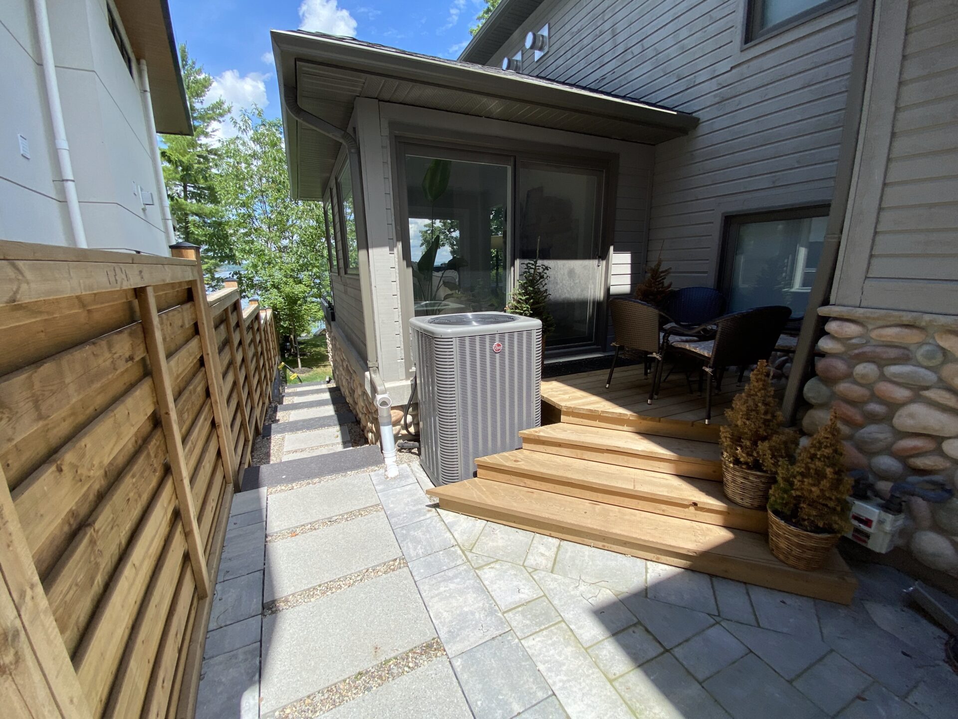 A modern house features a small deck with seating, surrounded by a wooden fence. A central air conditioning unit is visible.