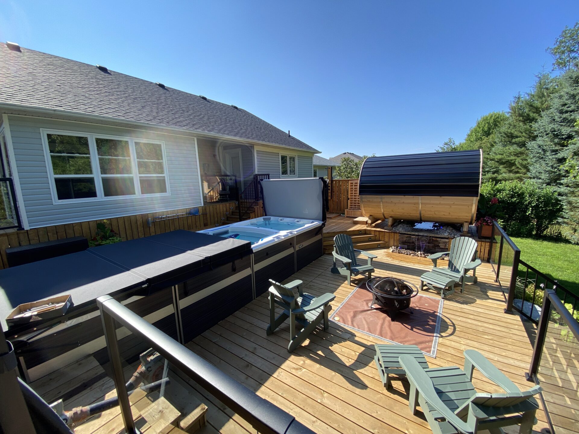 A spacious wooden deck features a hot tub, fire pit, seating area, and covered outdoor structure, surrounded by lush greenery on a sunny day.