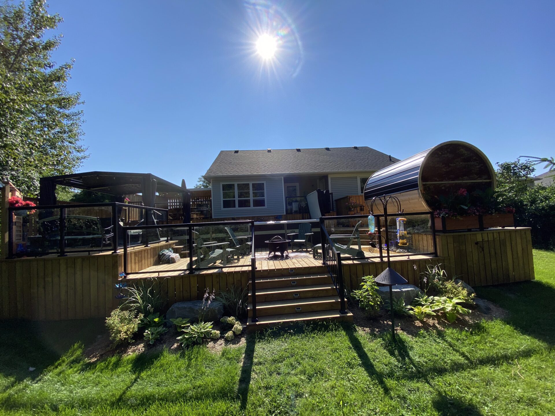 A sunny day reveals a backyard with wooden decking, stairs, seating, and a barrel sauna, surrounded by a well-maintained lawn and trees.