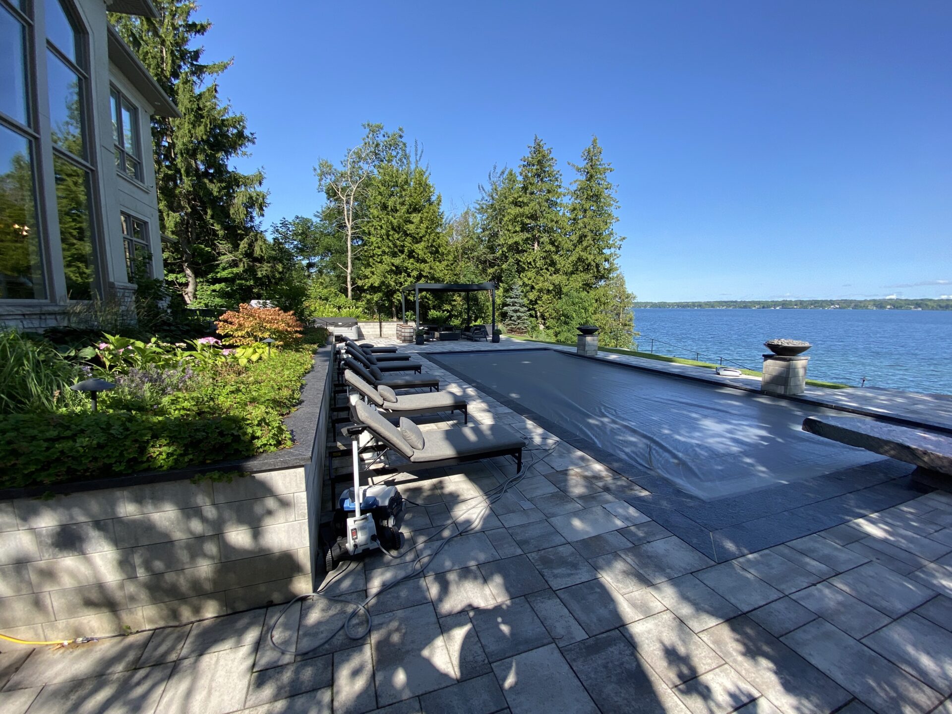 Poolside area with covered pool, lounge chairs, and lush greenery. Scenic view of calm lake and distant land under a clear blue sky.