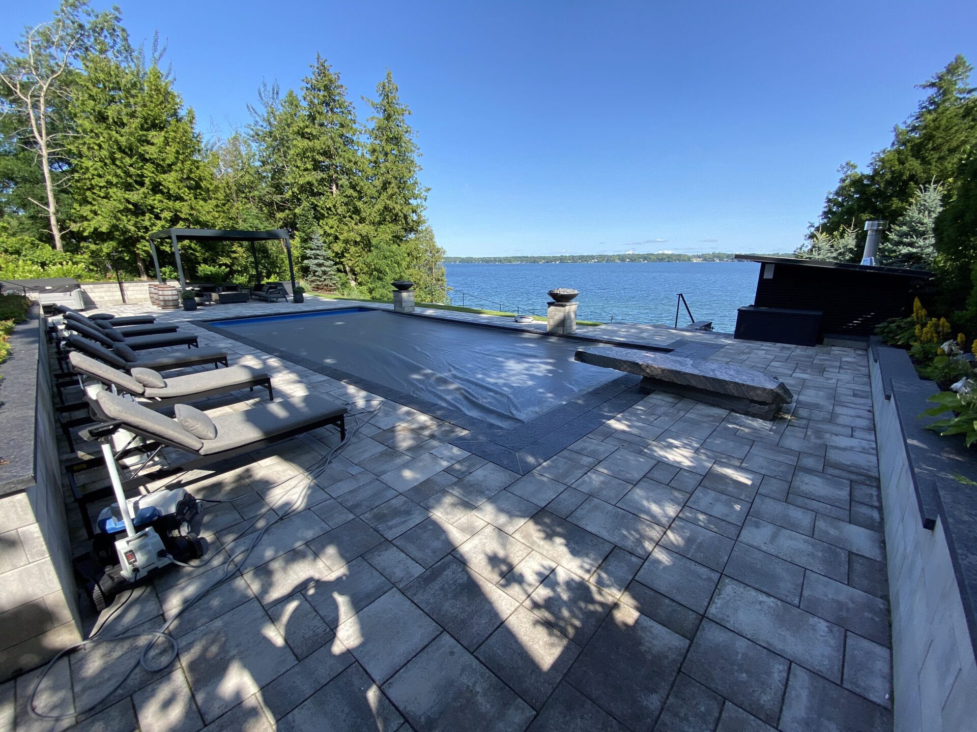 Lakeside patio with covered pool, sun loungers, and lush trees. Clear sky and calm water create a serene, relaxing outdoor environment.