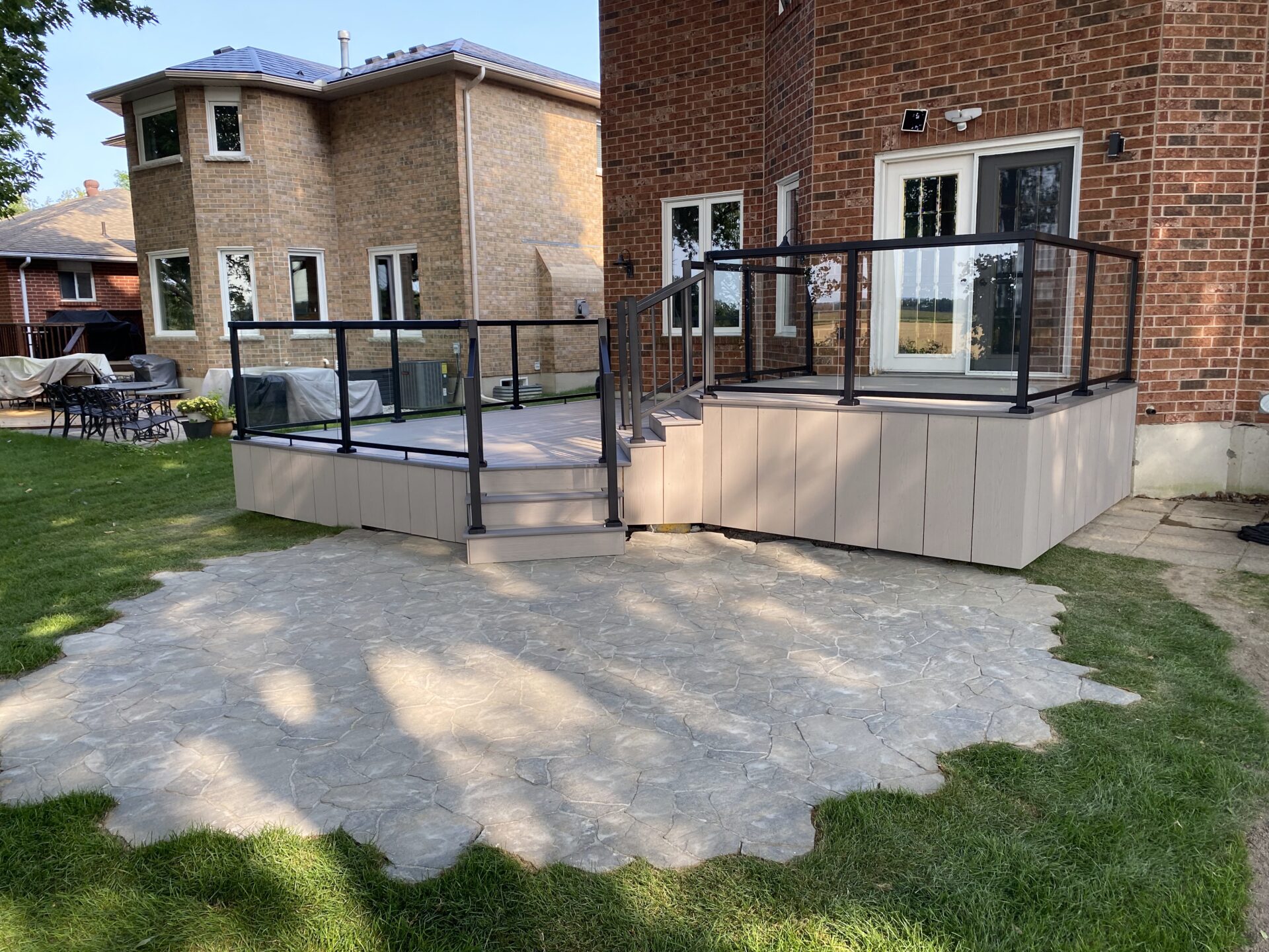 A modern wooden deck with glass railings attached to a brick house, overlooking a stone patio and grassy yard.