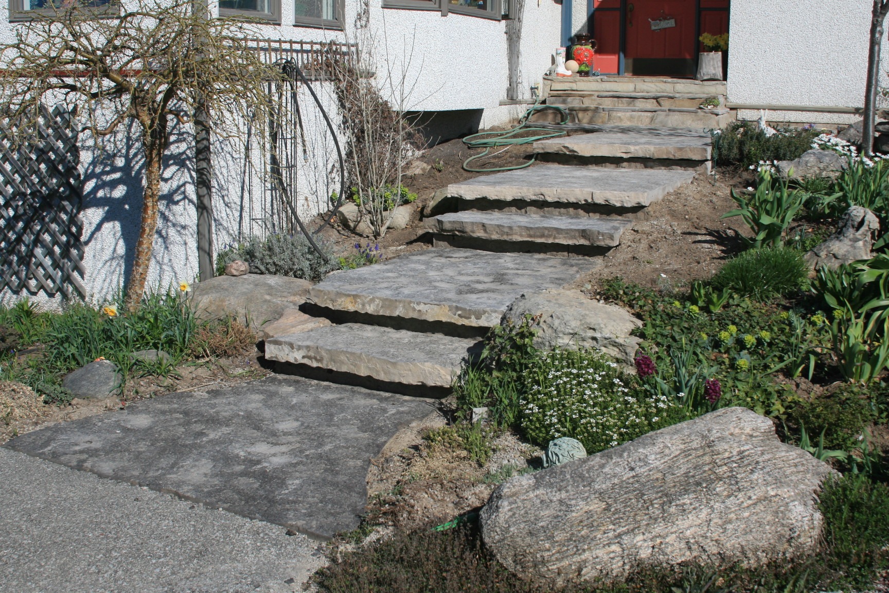 Stone steps lead to a house entrance, surrounded by rocks and garden plants. A coiled hose is visible near the door.