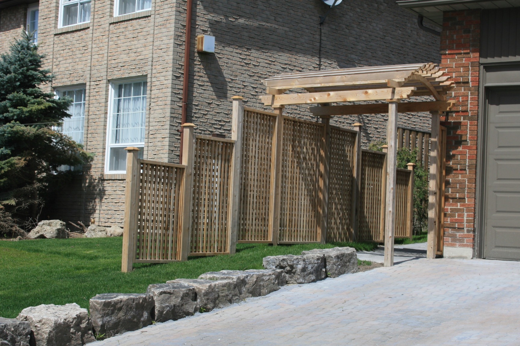 A residential outdoor space features a wooden trellis and fence, stone border, brick house facade, windows, and green lawn under a clear sky.